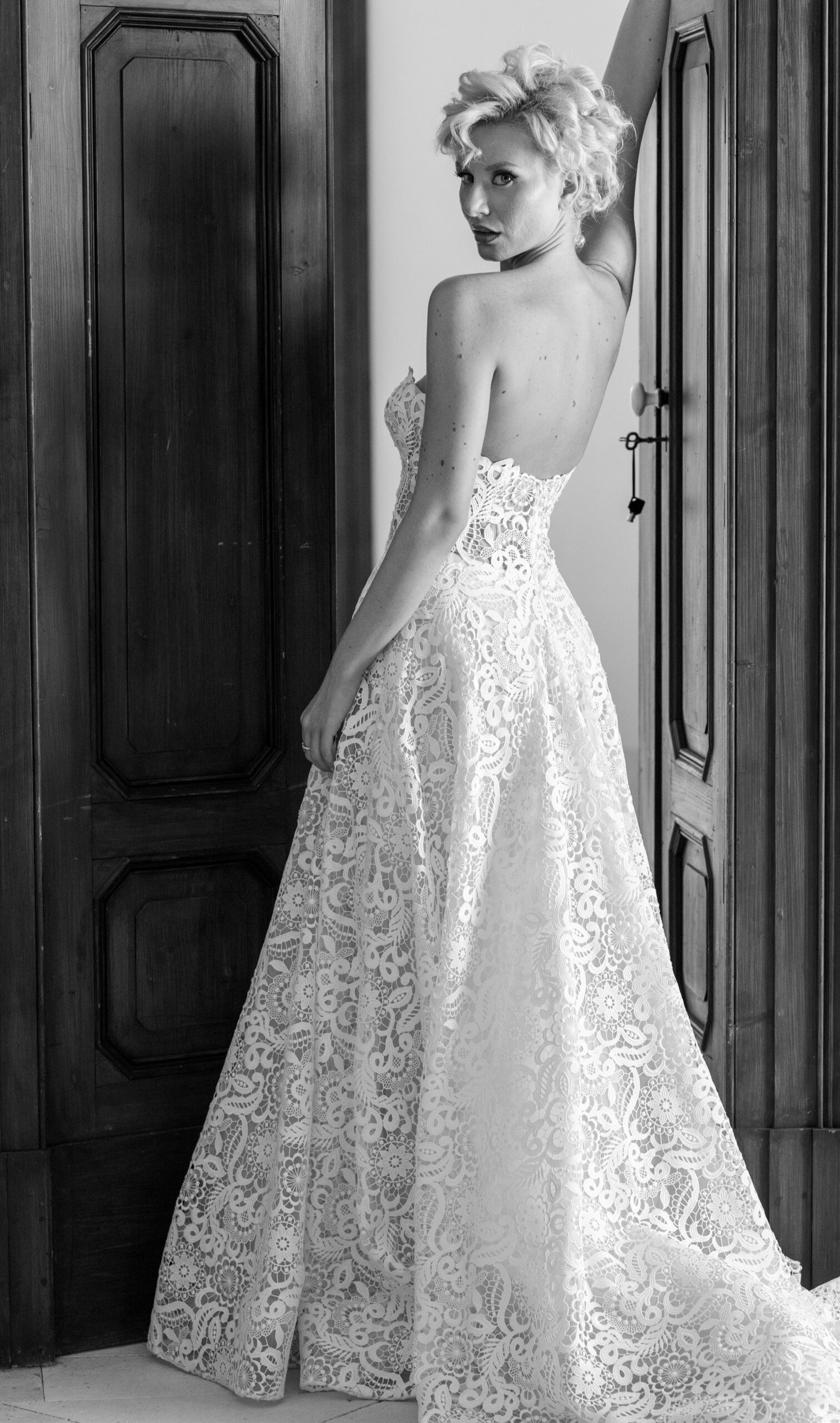 A woman with short, curly blonde hair wears a strapless, lace wedding gown, standing between two wooden doors and looking over her shoulder in a black and white photograph.