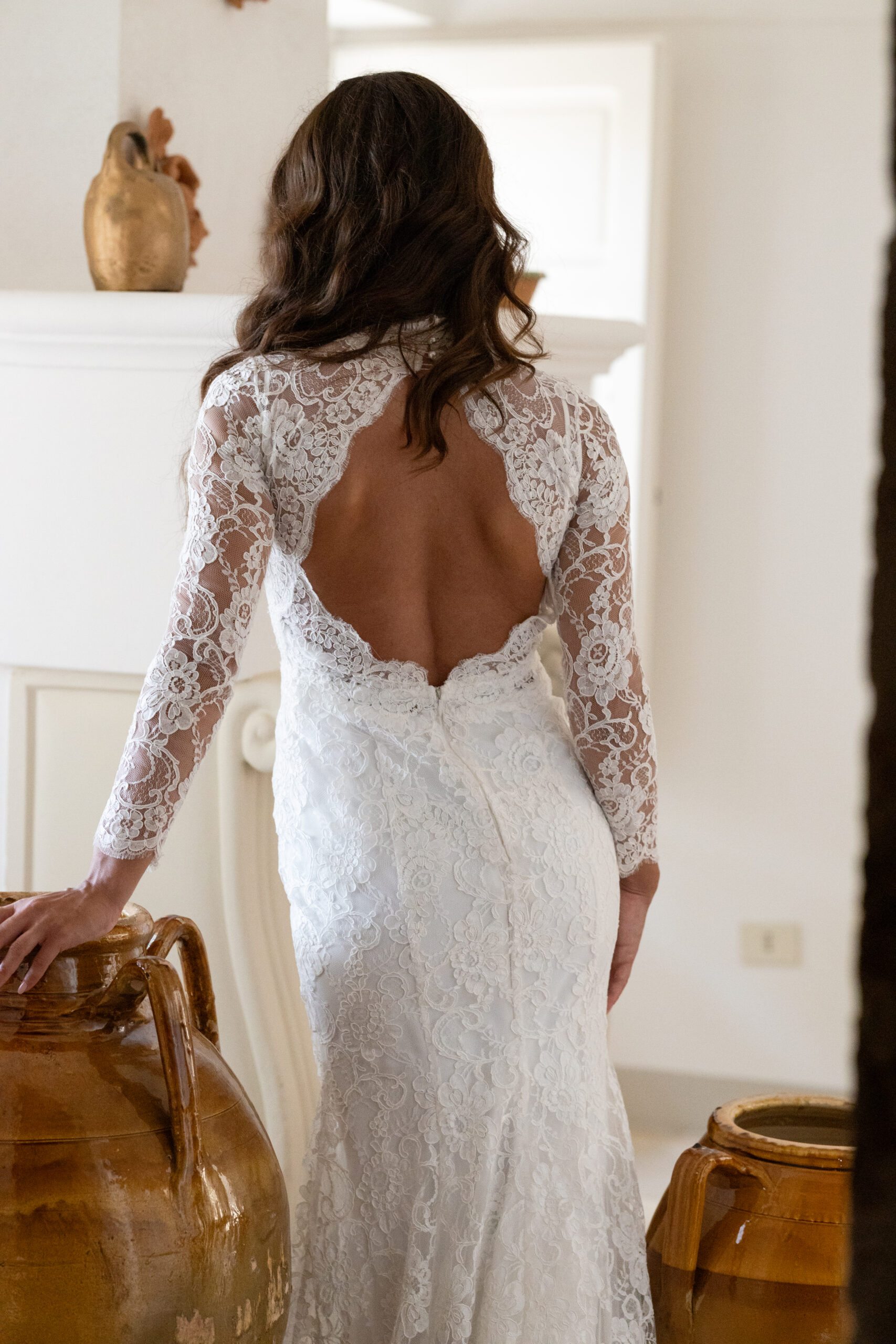 A woman in a white lace wedding dress with an open back stands indoors, her back to the camera, with her hand resting on a large brown ceramic vase.