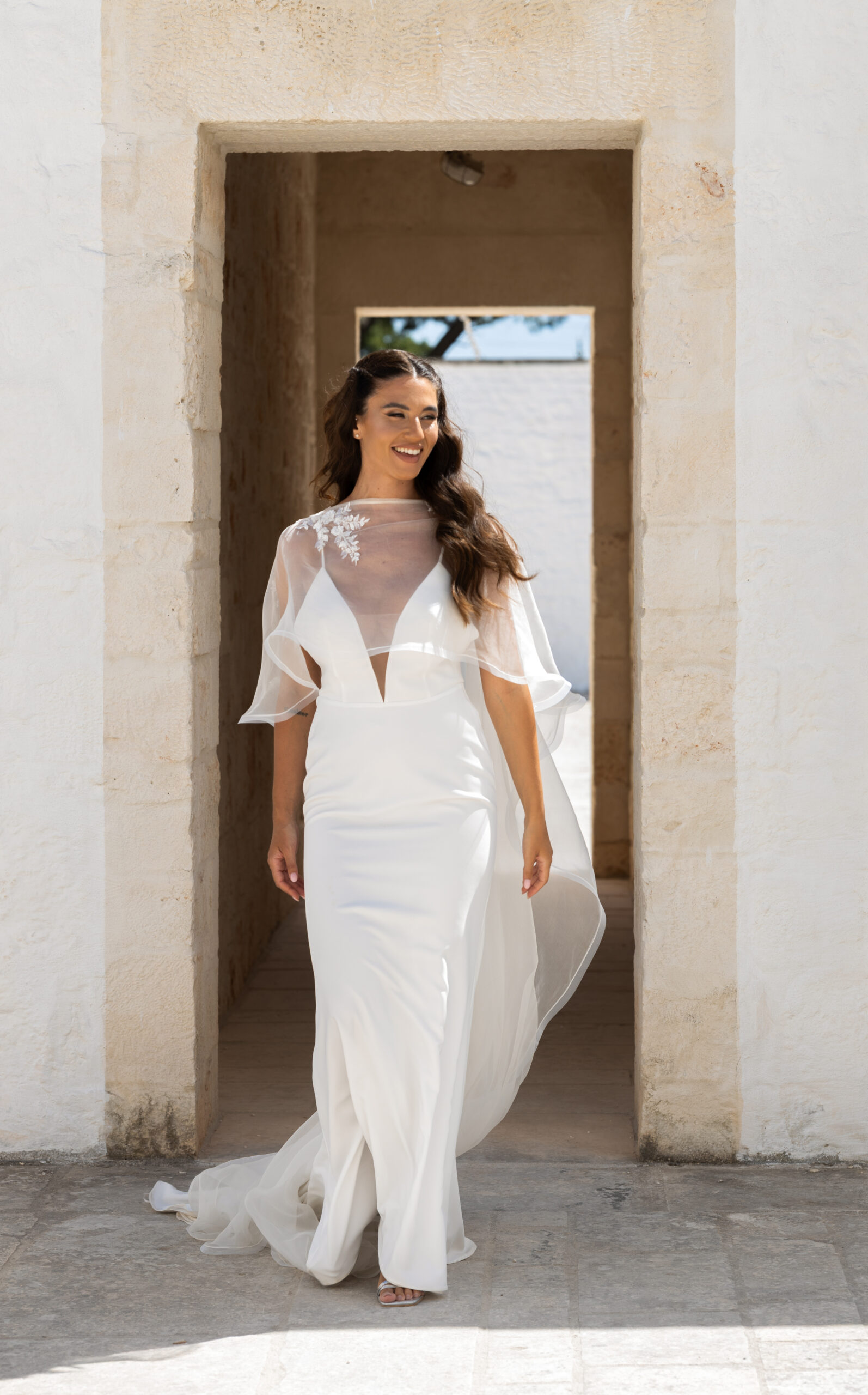 A woman in a white wedding dress with sheer, floral accents walks through a stone archway, smiling and looking to the side, with natural sunlight illuminating the scene.