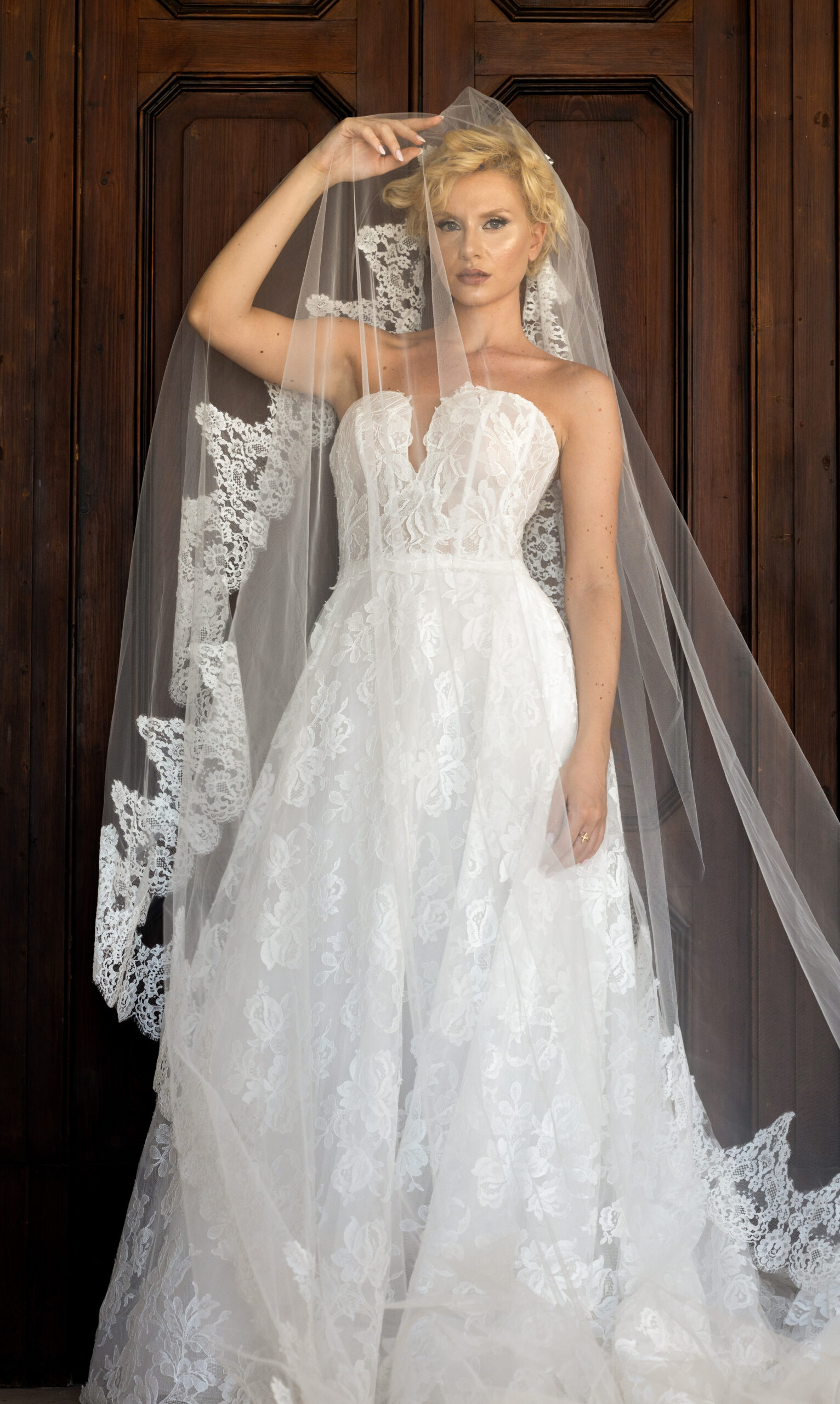 A woman in a strapless white lace wedding dress and long lace veil stands in front of a wooden door, looking directly at the camera while holding part of her veil above her head.