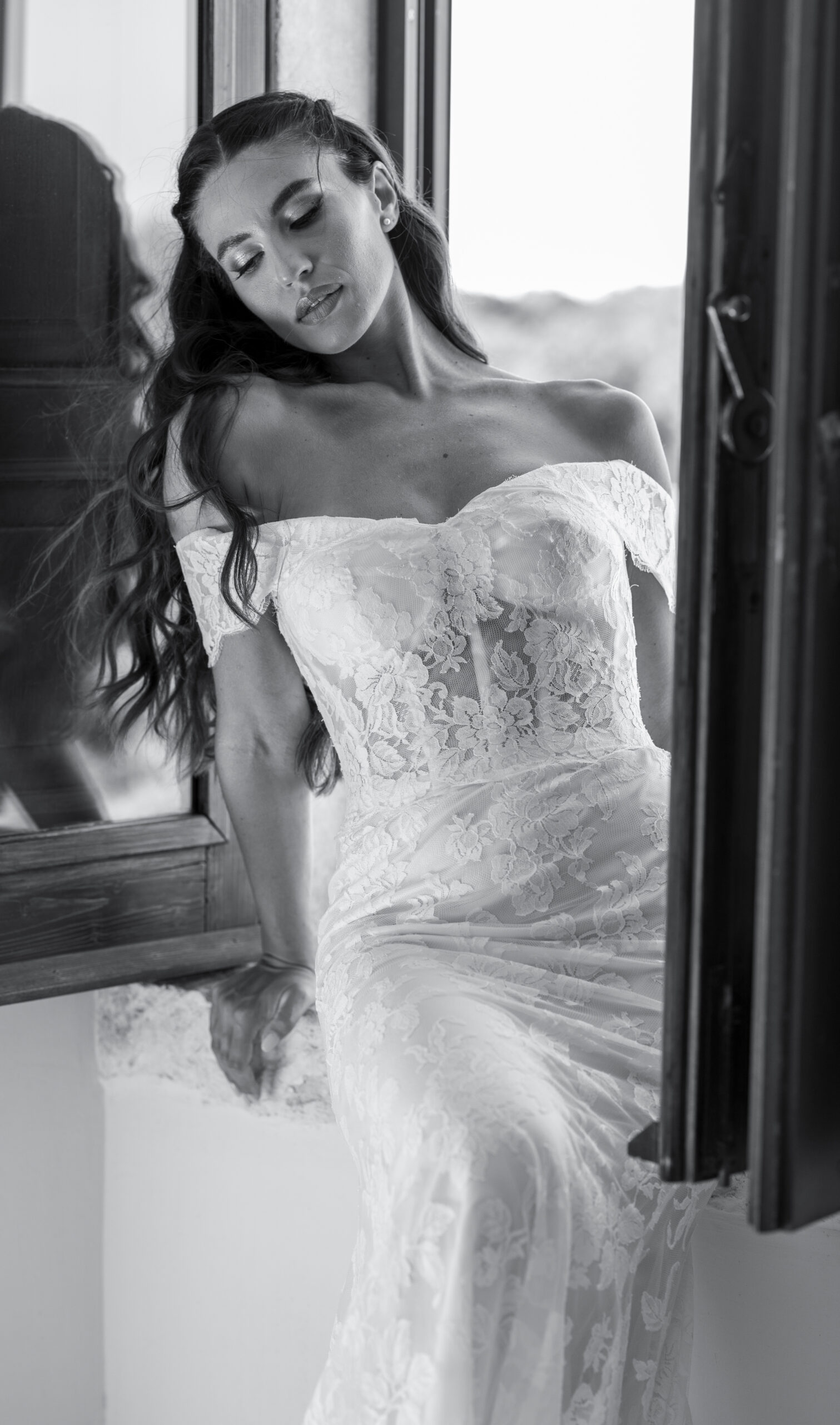 A woman in an off-the-shoulder lace wedding dress sits on a windowsill, eyes closed, with long wavy hair cascading over her shoulder. The photo is black and white, highlighting the delicate details of her gown.