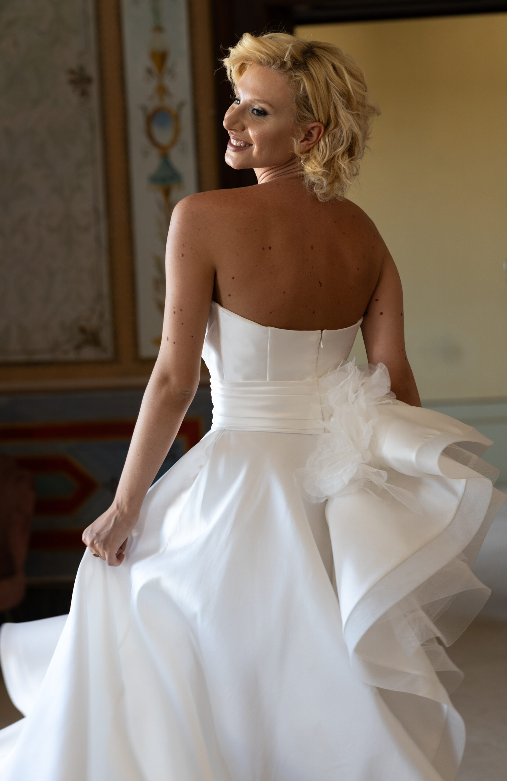 A smiling woman with short blonde hair wearing a strapless white wedding dress looks back over her shoulder, holding part of her dress as she stands indoors in a decorated room.