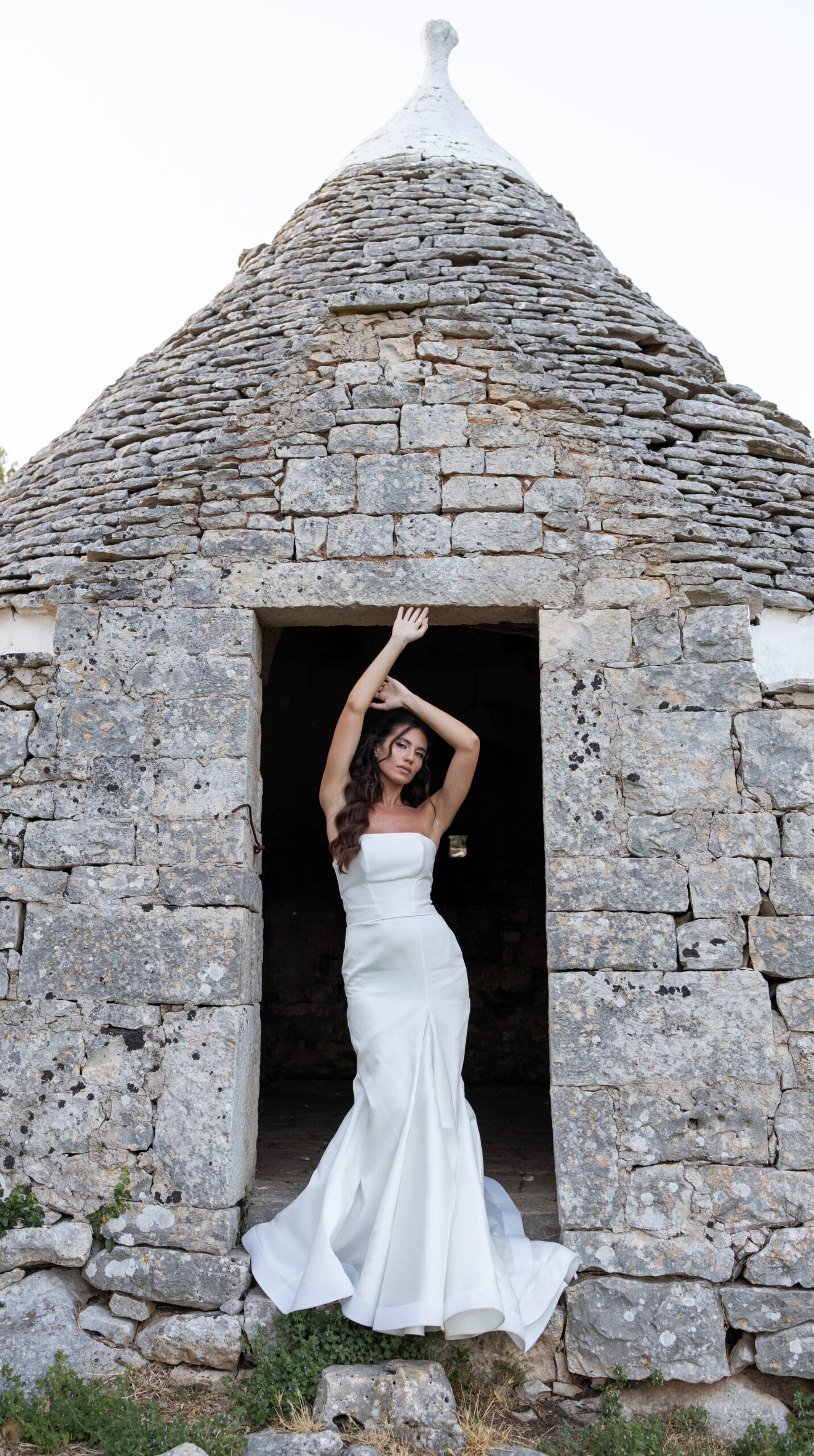A woman in a strapless white gown stands in the doorway of an old stone building with a conical roof, posing with her arms raised above her head.