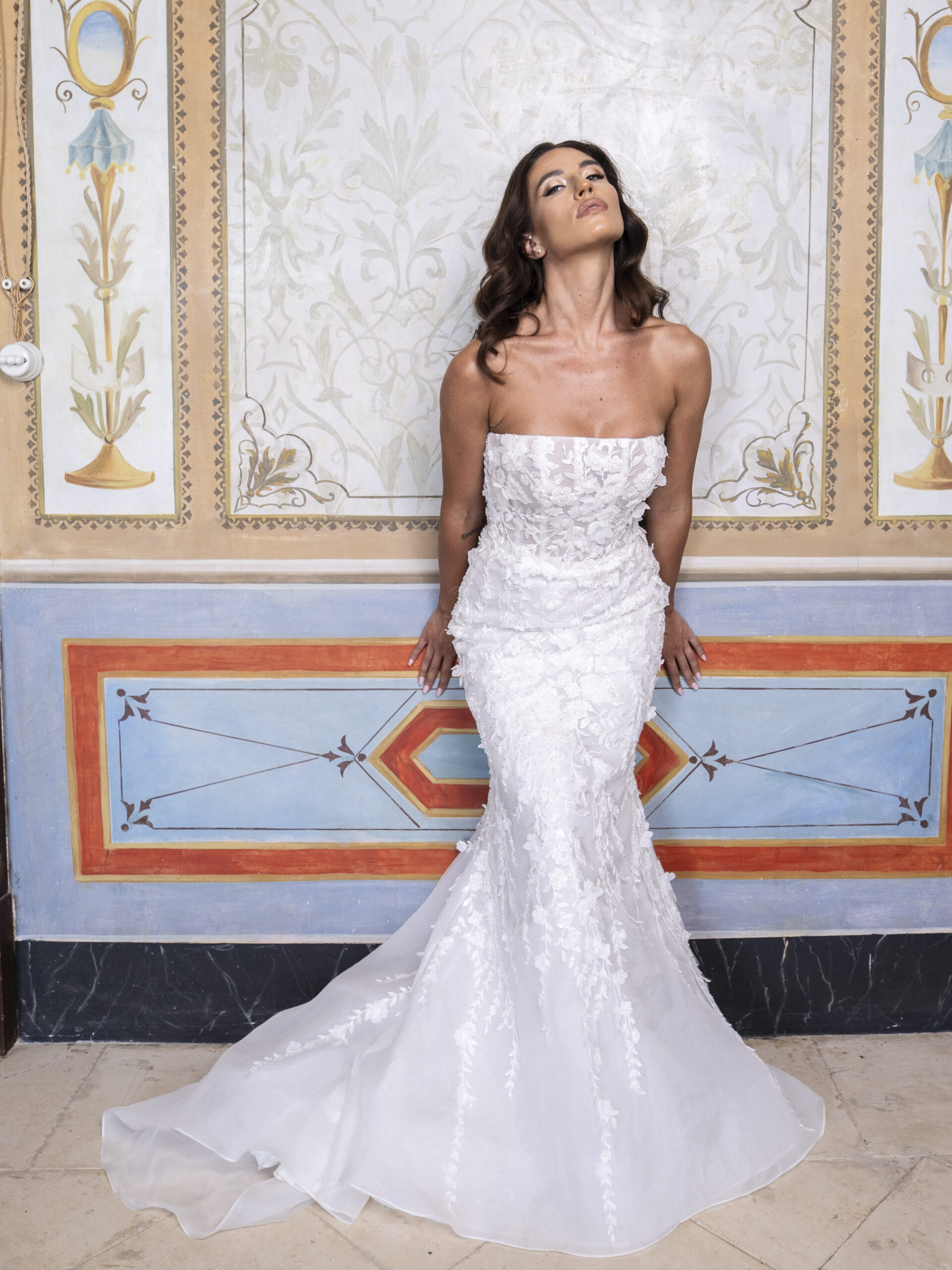 A woman in a strapless, fitted white lace wedding dress poses against an ornate, decorative wall with floral and geometric designs, looking upward with her eyes closed.