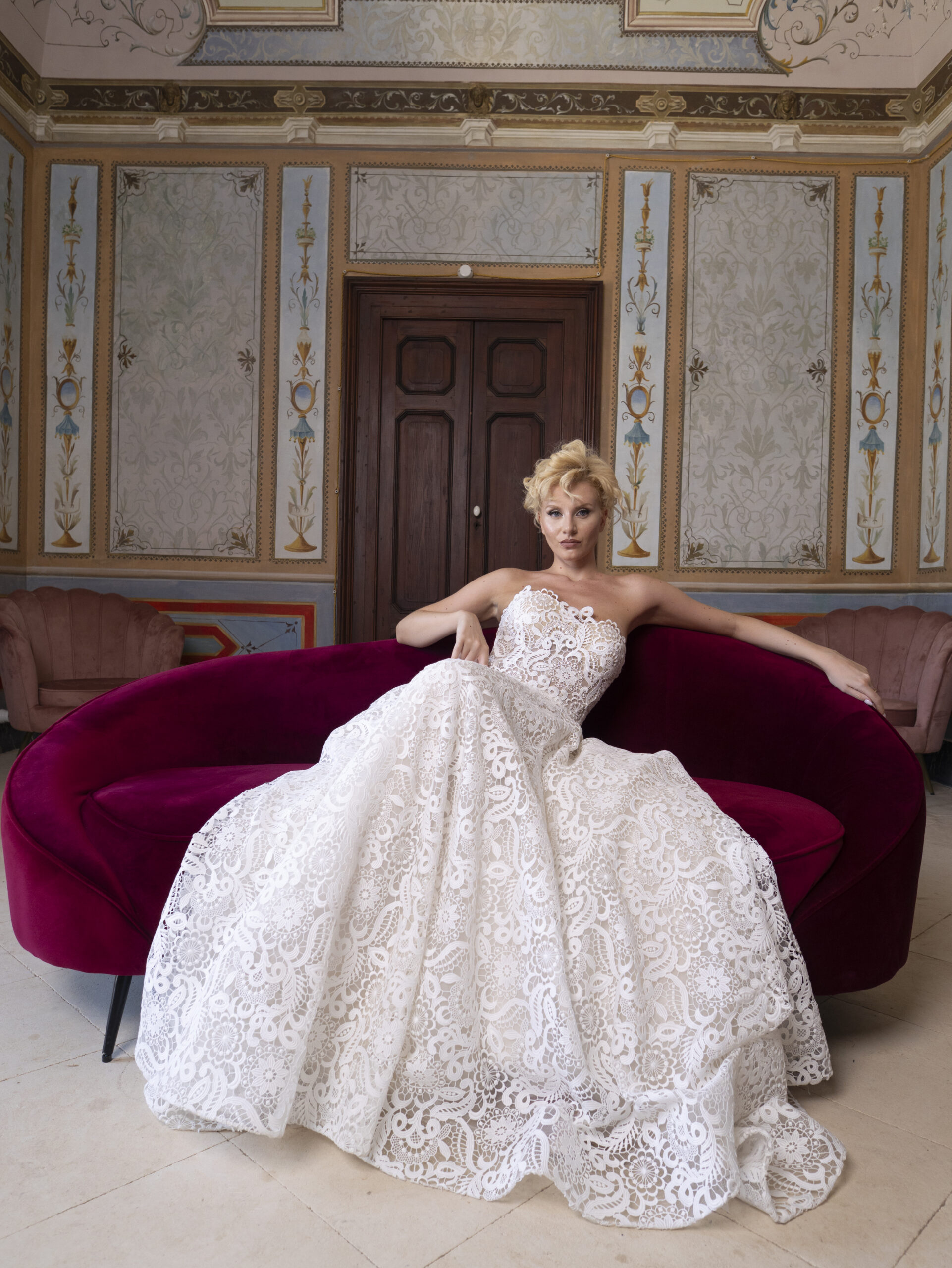 A woman in a white lace strapless gown sits on a curved red velvet sofa in an ornate room with decorative walls and a wooden door behind her.