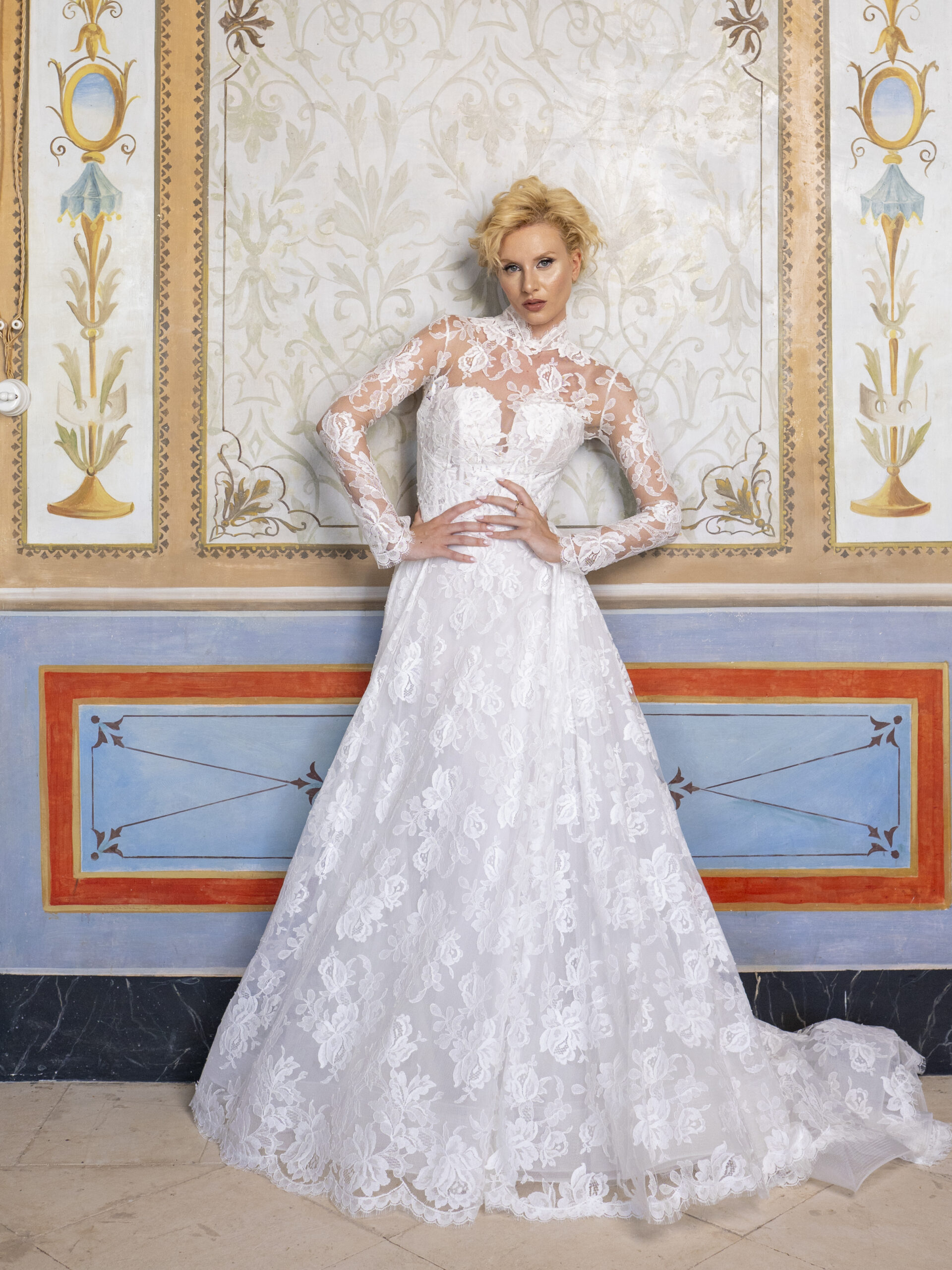 A woman in a long-sleeved, lace white wedding dress stands against an ornate, decorated wall with floral patterns and colorful panels.