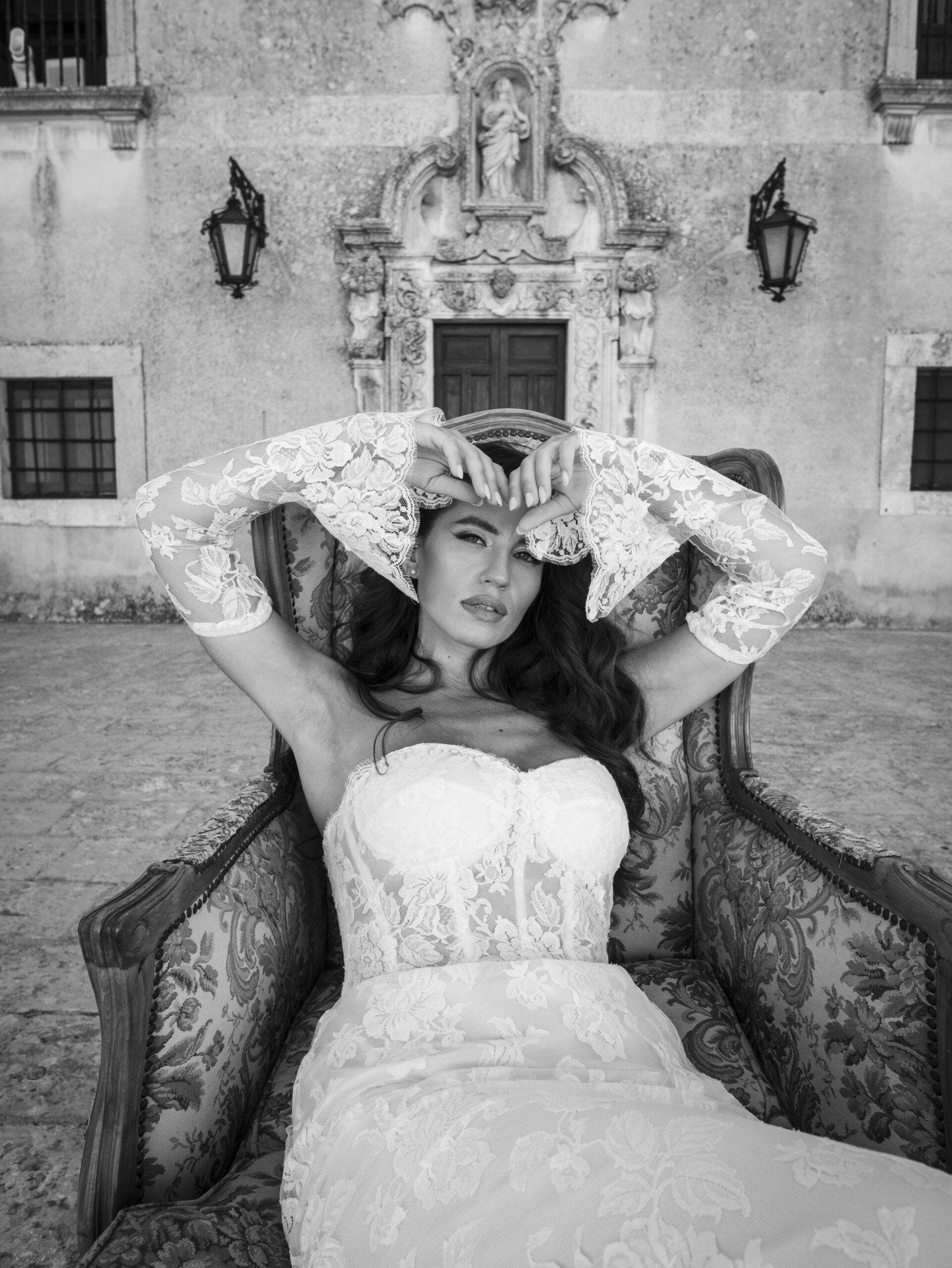 A woman in a strapless lace wedding dress lounges on an ornate chair outdoors, forming a heart shape with her gloved hands. The background features an old, decorative building façade. The image is black and white.