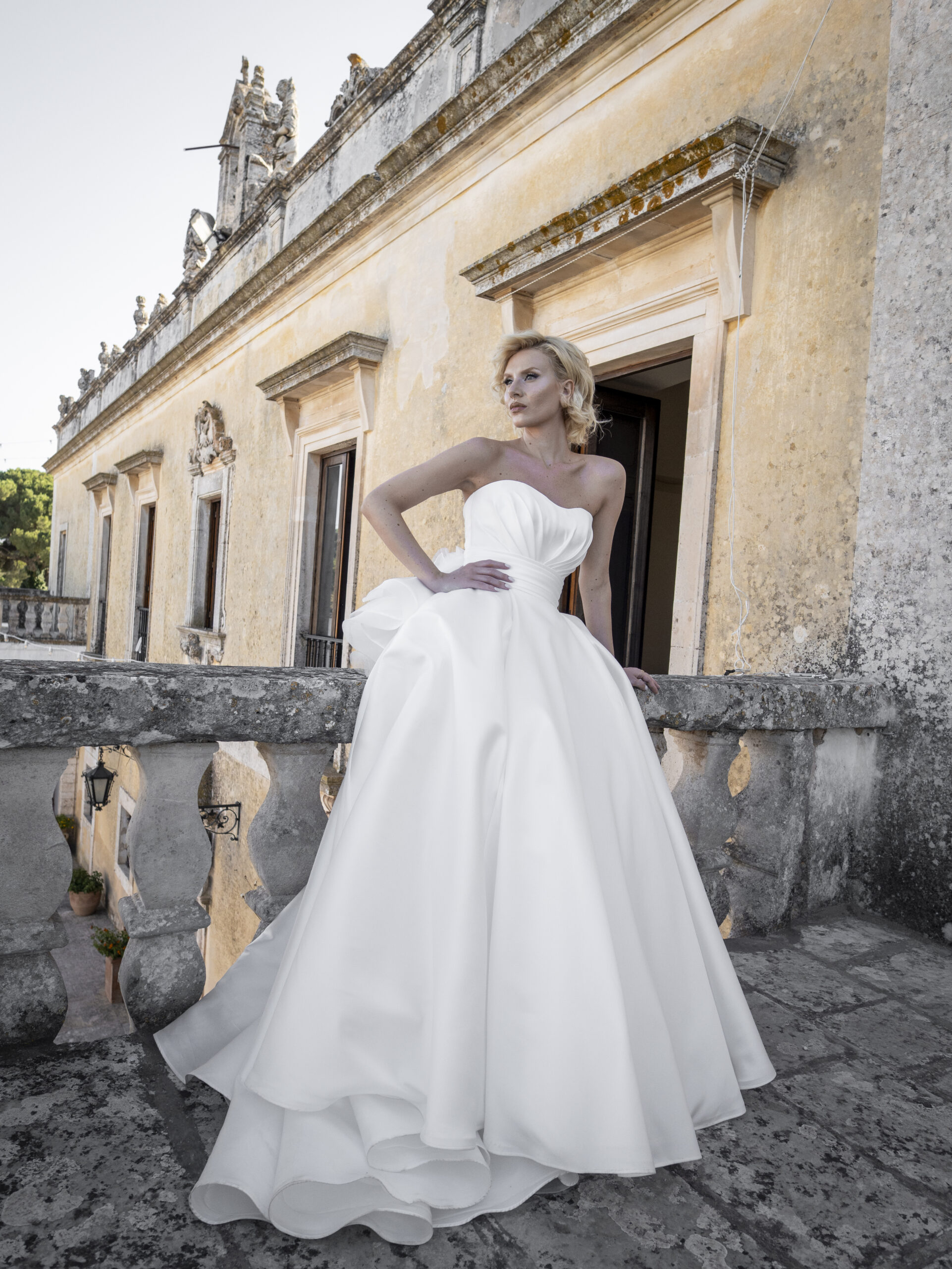 A woman in a strapless white wedding gown stands on a stone balcony, leaning against the balustrade, with an old, ornate building in the background.