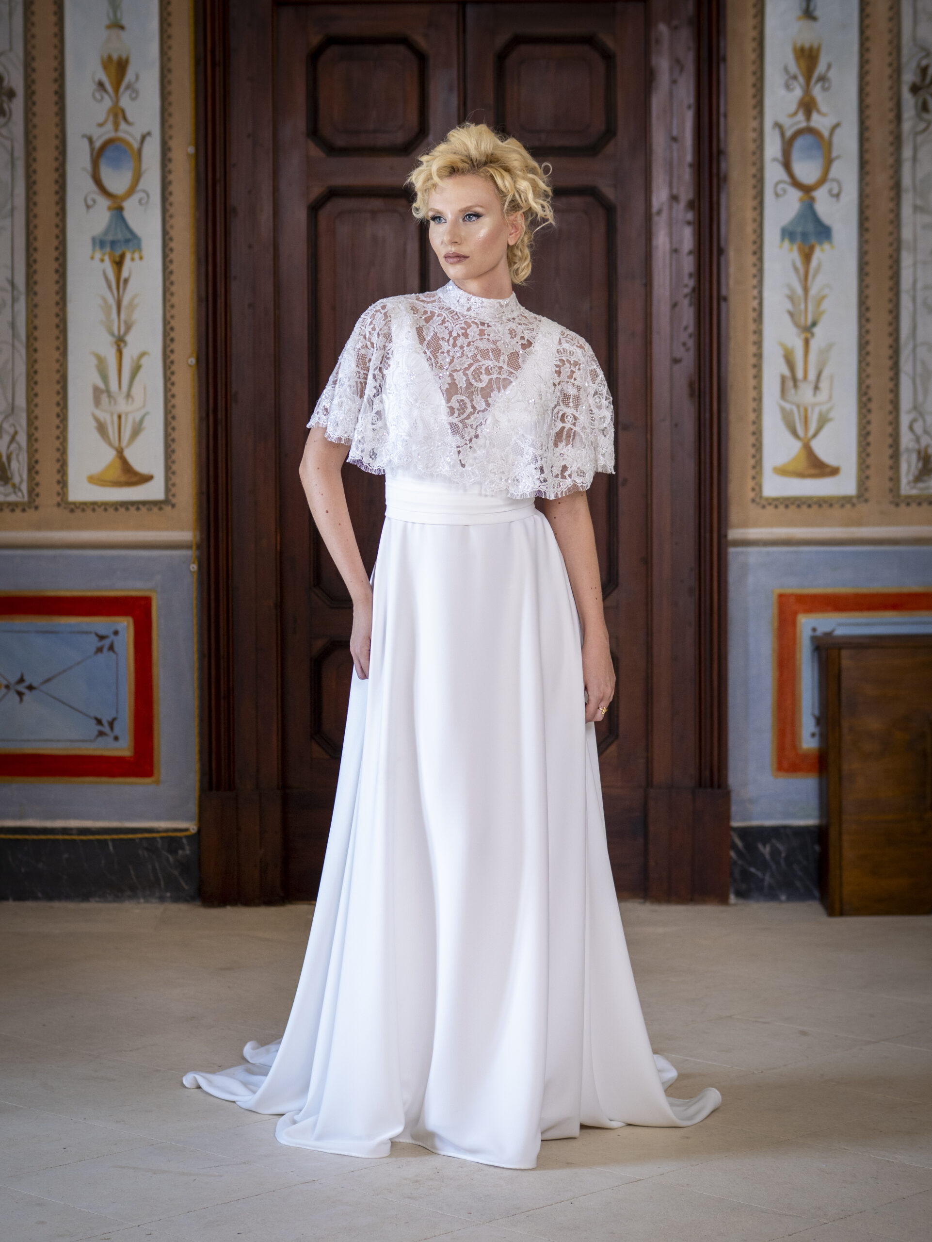 A woman in an elegant white wedding dress with a lace top and flowing skirt stands indoors in front of ornate, decorative walls and a wooden door.