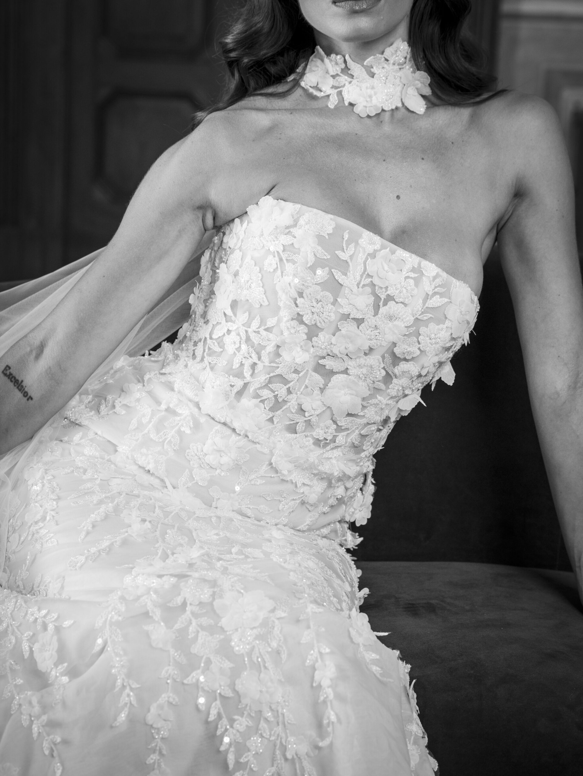 A black and white photo of a woman wearing a strapless, floral lace wedding dress, seated with one arm resting on her lap. She also wears a floral choker and has a visible tattoo on her arm.