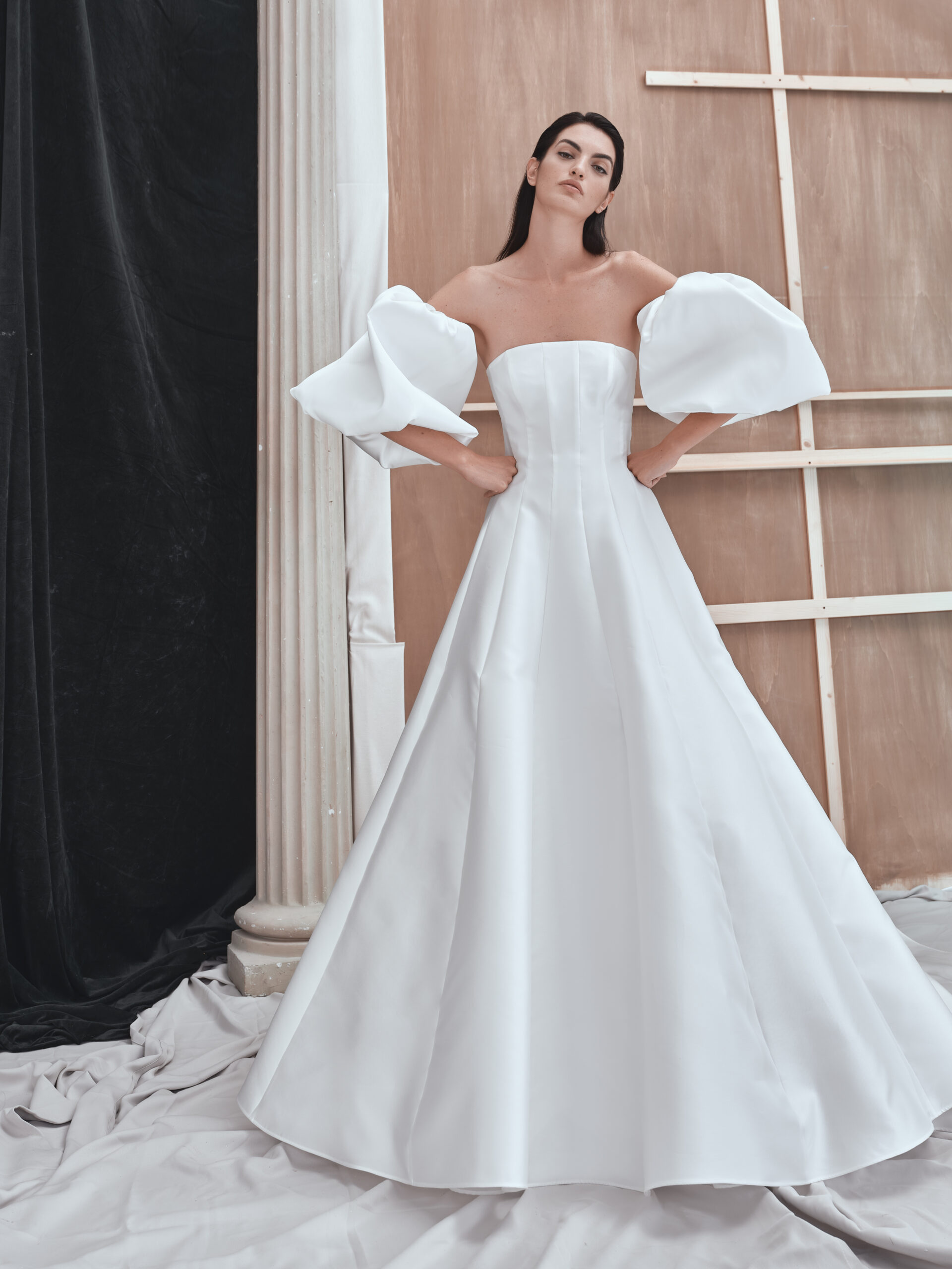 A woman wearing an elegant off-the-shoulder white gown with voluminous puff sleeves stands with her hands on her hips in front of a column and unfinished wooden backdrop.