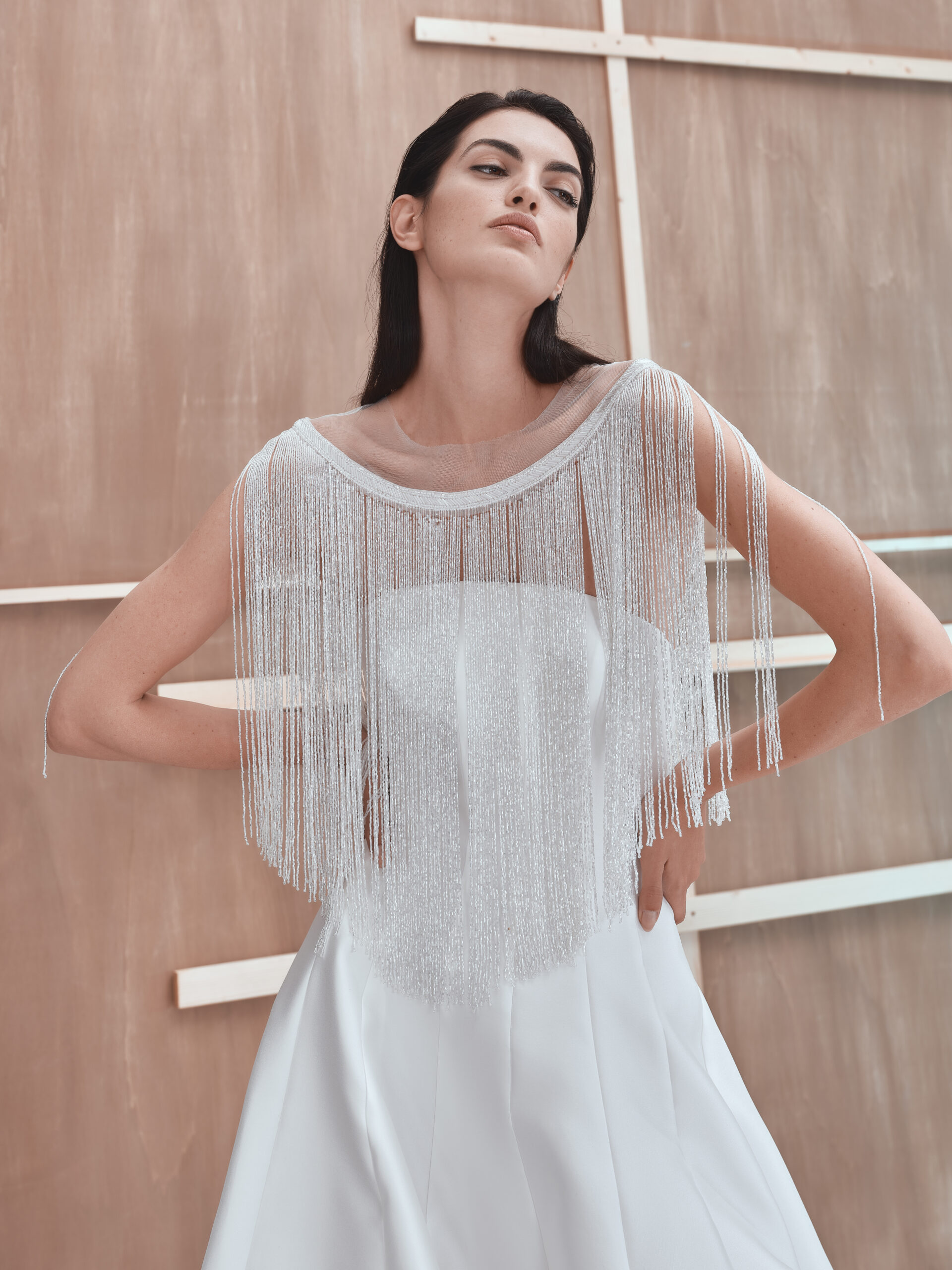 A woman in a white dress with a sheer, beaded fringe cape stands confidently with hands on hips against a wooden panel background with white geometric accents.