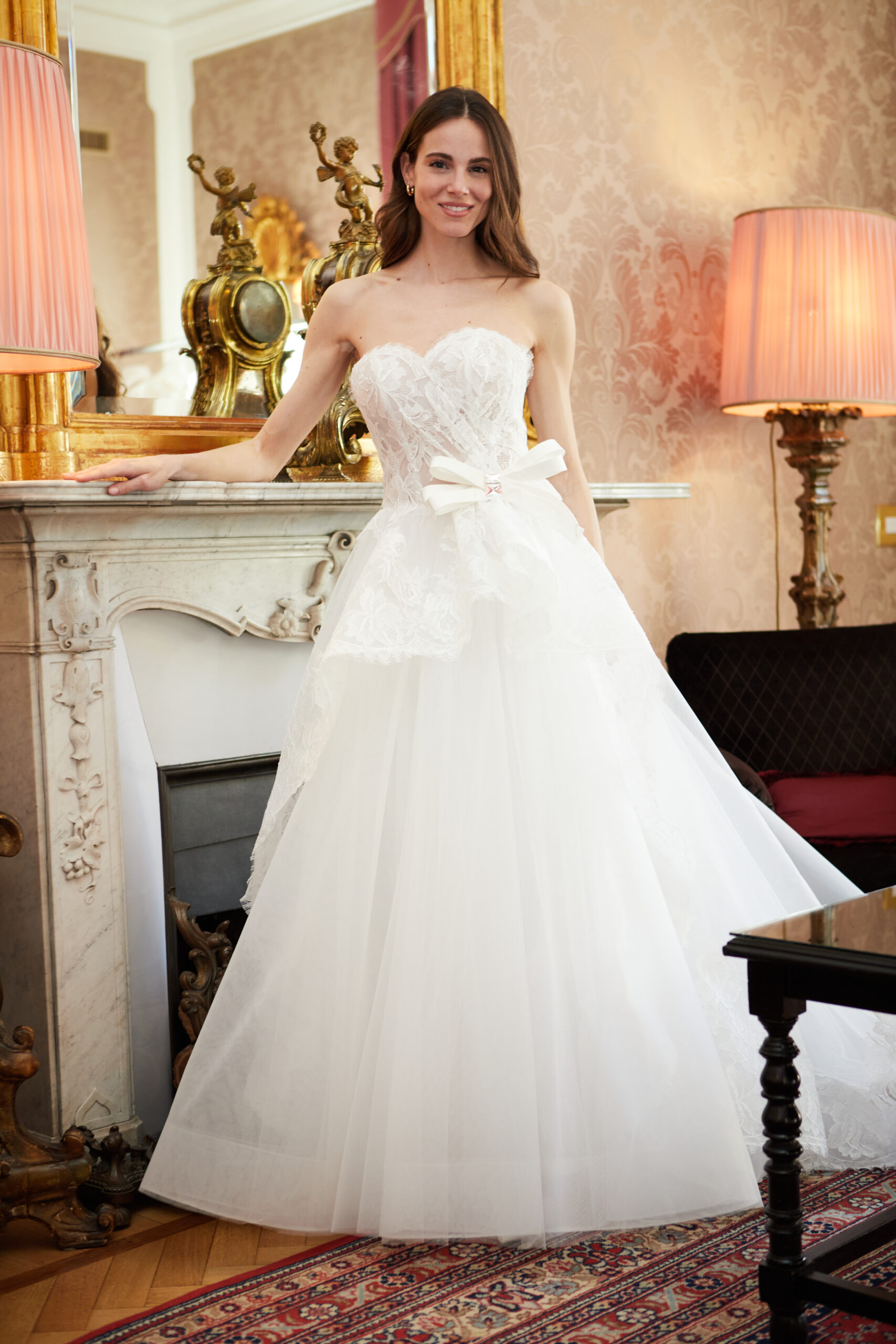 A woman in a strapless, lace, white wedding gown with a large bow stands by a marble fireplace in an elegant room with ornate decor and two tall, glowing lamps.