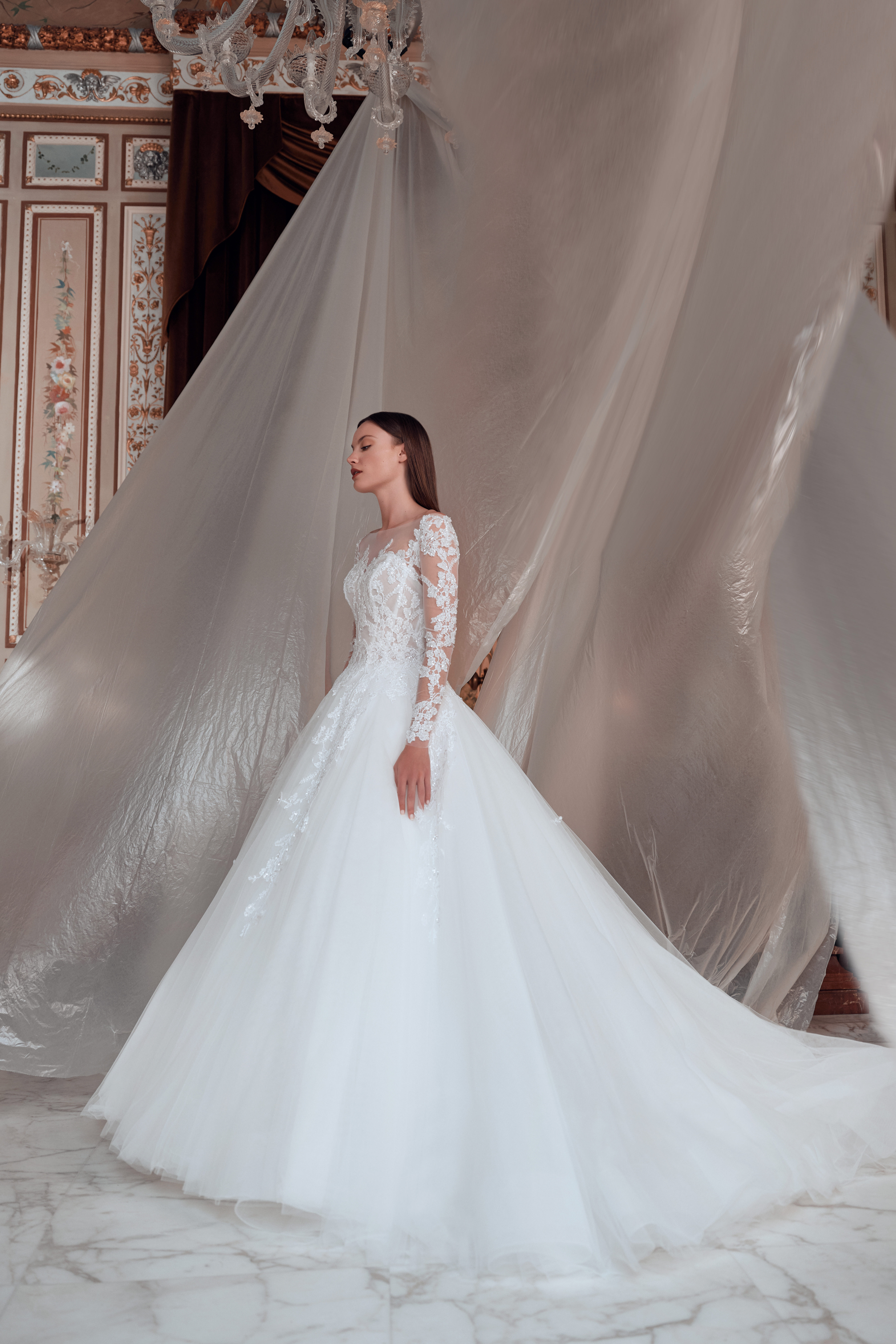 A woman in a long-sleeved, lace wedding dress stands indoors on marble flooring, with elegant, ornate walls and flowing sheer fabric draped behind her, creating a dramatic and sophisticated atmosphere.