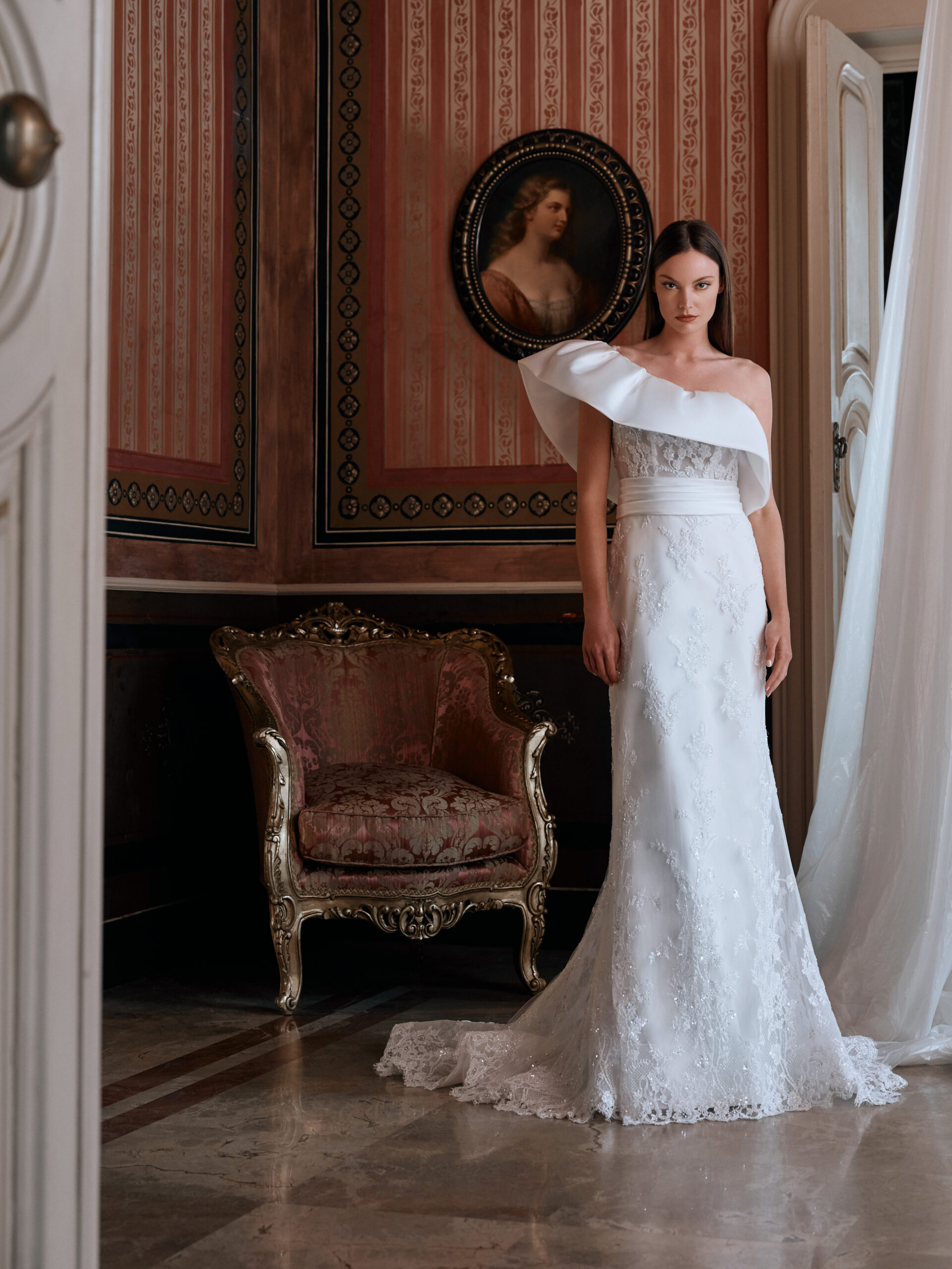 A woman in an elegant white, one-shoulder wedding gown stands in a vintage room with ornate wallpaper, a gold-trimmed antique chair, and a classical portrait hanging on the wall behind her.