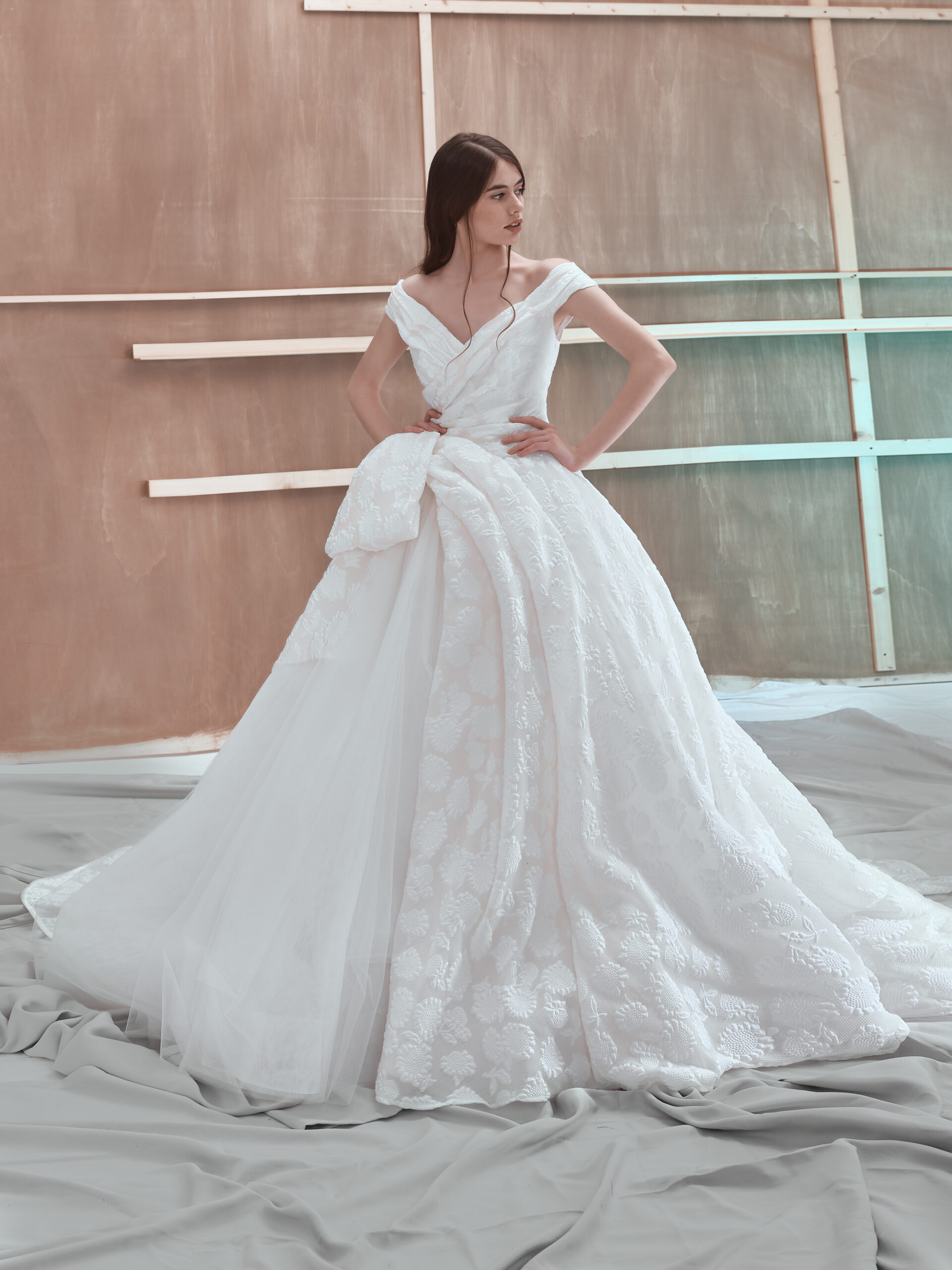 A woman poses in a white, off-the-shoulder wedding gown with a full, textured skirt and a large bow detail, standing indoors in front of an unfinished wooden wall.
