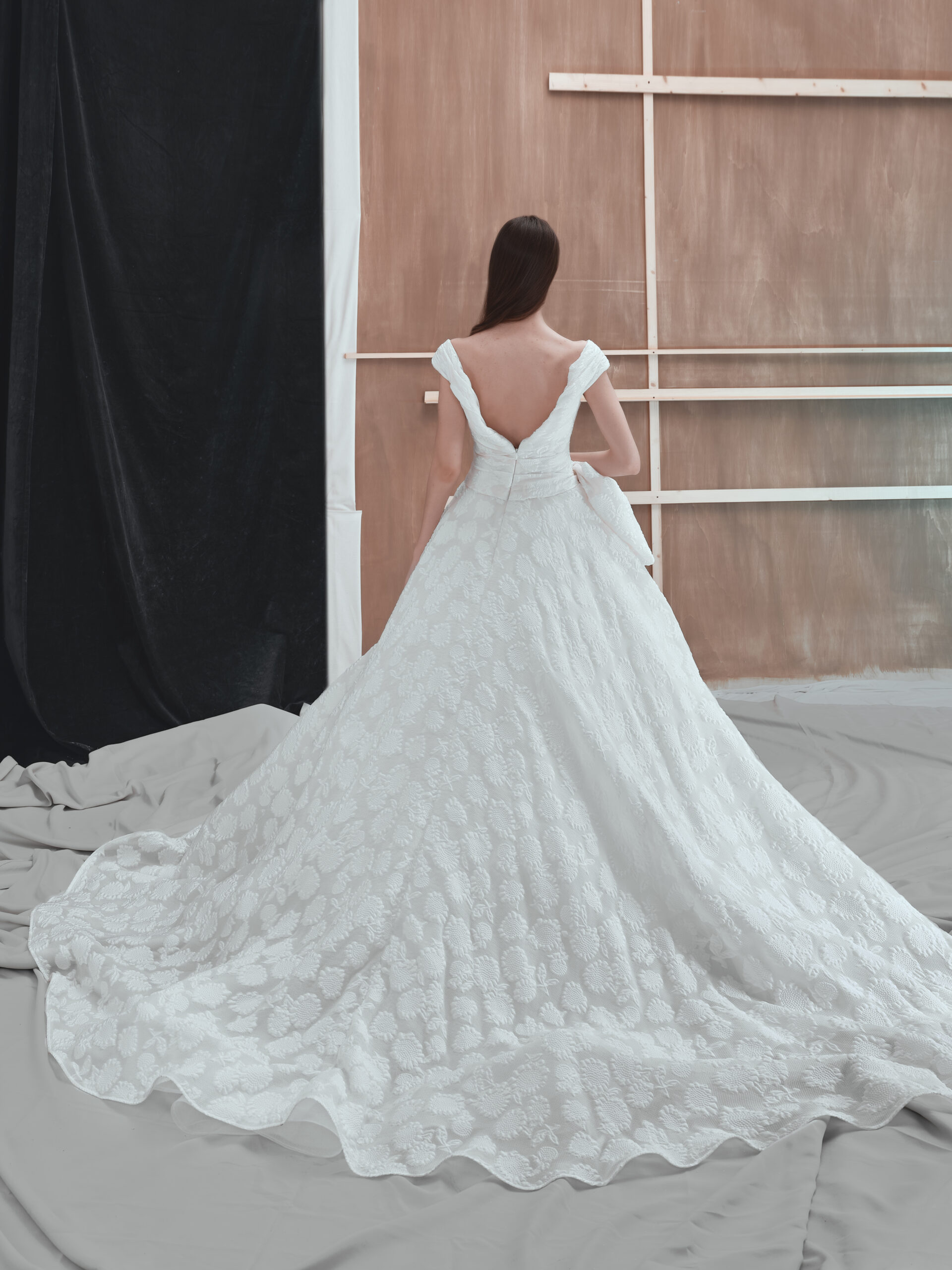 A woman with long brown hair stands with her back to the camera, wearing an off-shoulder, white lace gown with a long, elegant train. She is in a studio with draped gray fabric and a wooden backdrop.