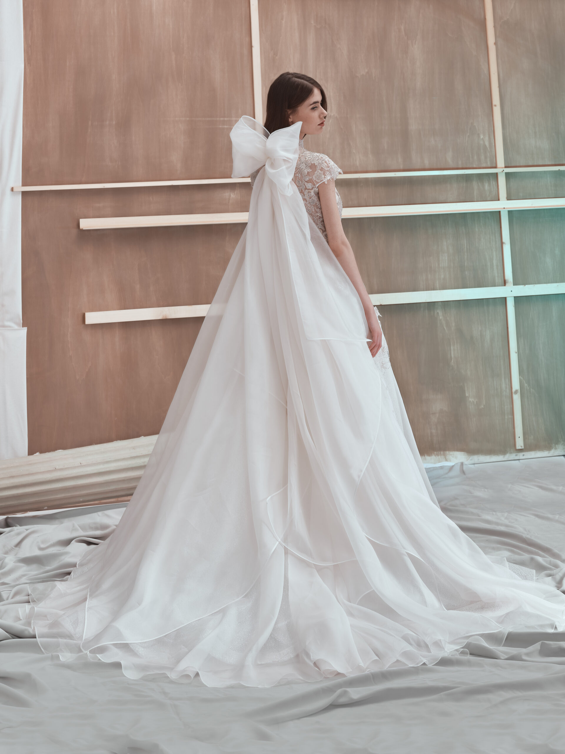 A woman in a white wedding dress with a large bow on the back stands on a draped fabric floor, facing away in front of a wooden, unfinished wall.