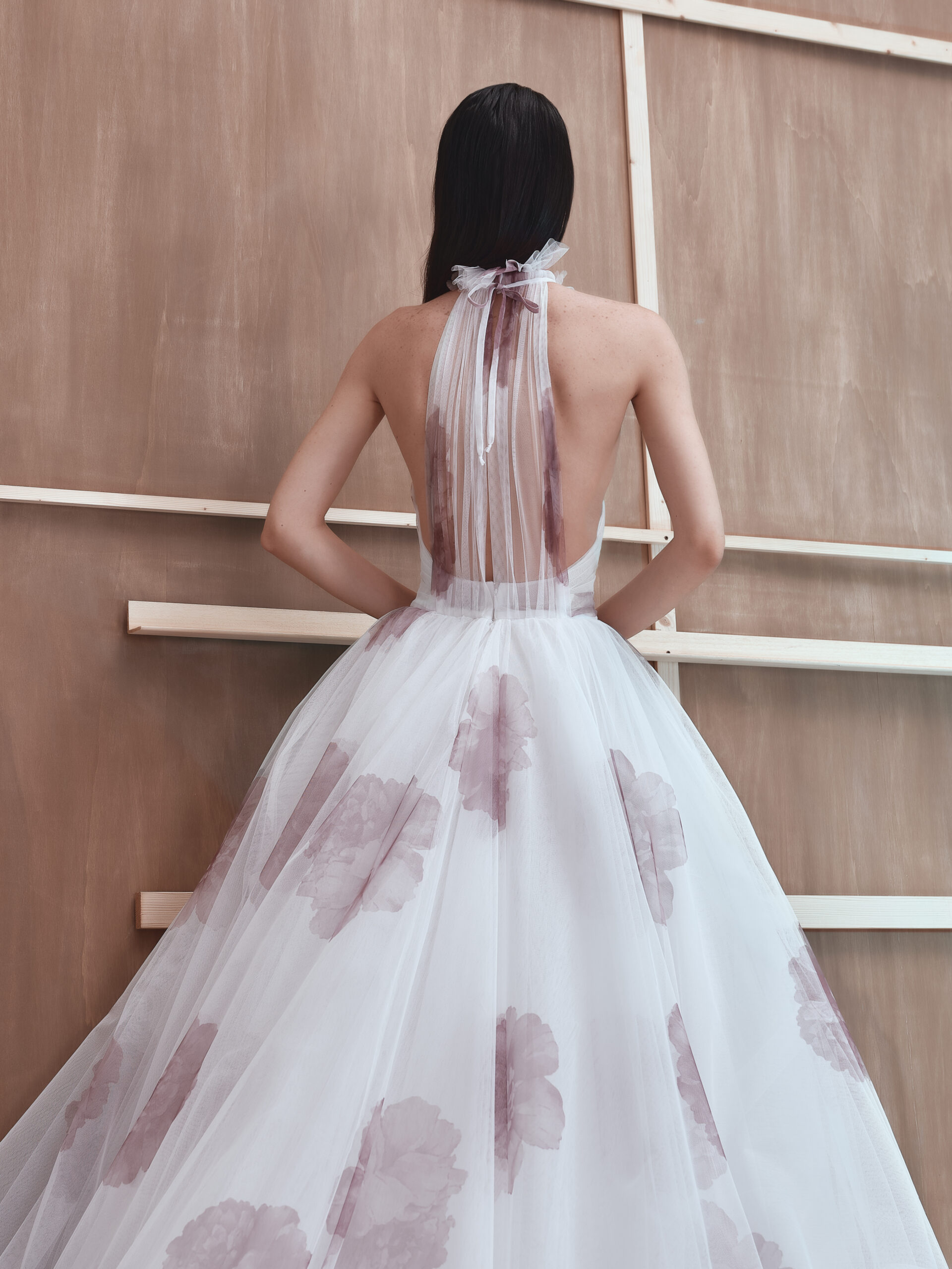 A woman stands with her back to the camera, wearing a white halter-neck gown with a tulle skirt decorated with large, soft mauve floral prints. The background features wooden panels and framework.