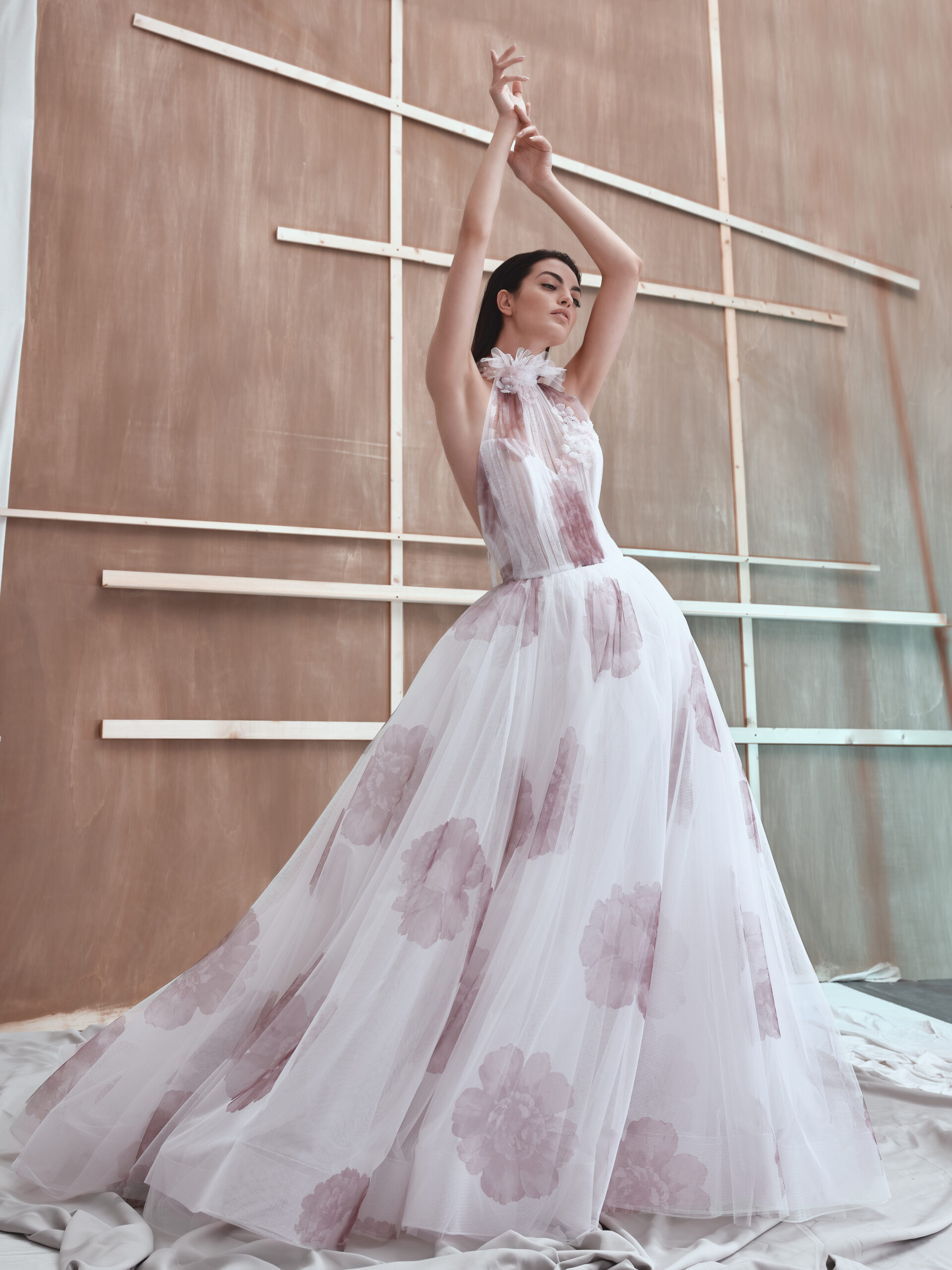 A woman in a flowing white gown with light pink floral patterns stands with her arms raised gracefully, posing in front of a wooden geometric backdrop. The dress is elegant and voluminous, and the scene is softly lit.
