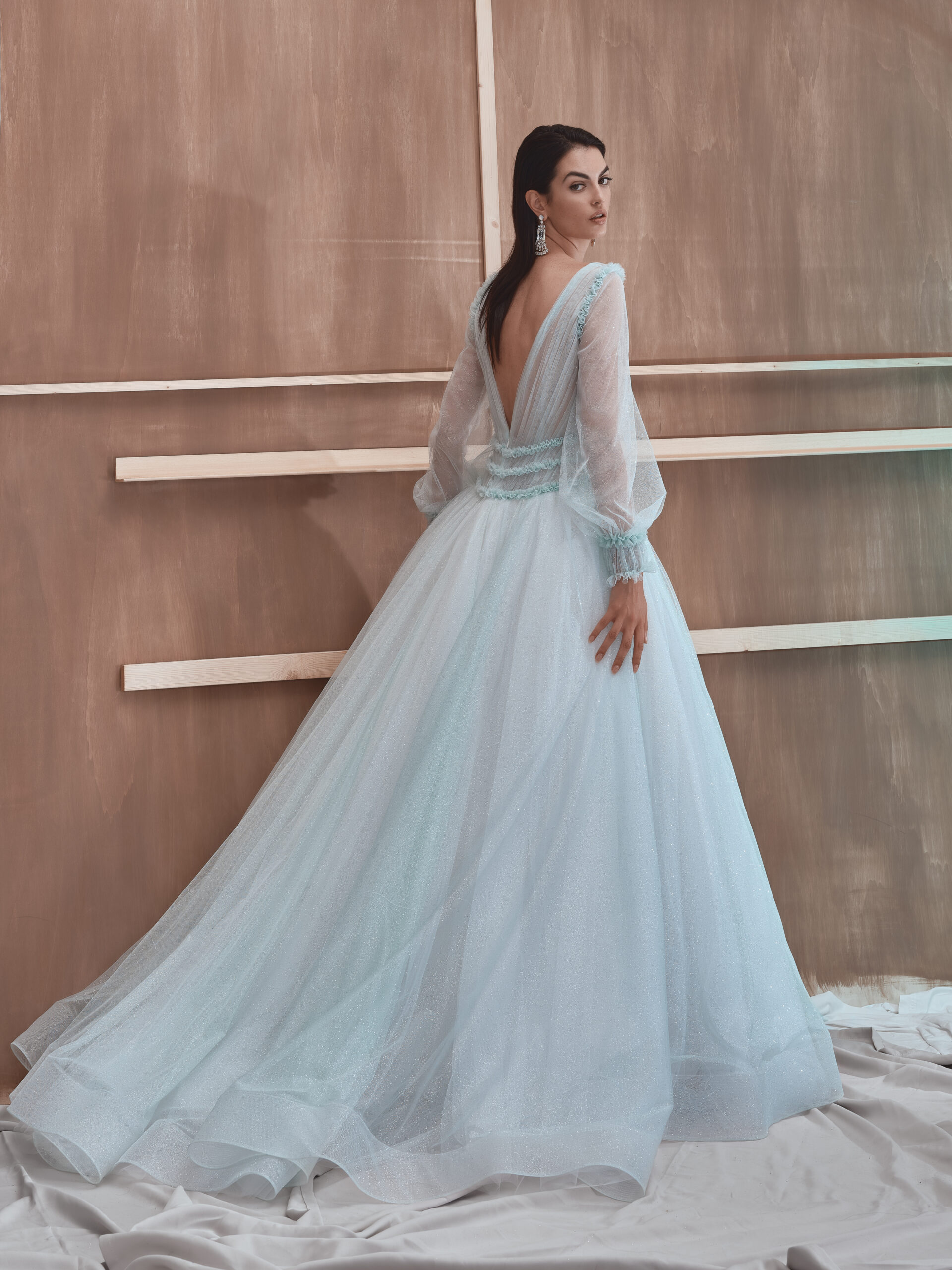 A woman in a flowing pale blue gown with sheer long sleeves poses with her back to the camera, revealing a deep V-back. She stands in front of a wooden textured wall with horizontal white accents.