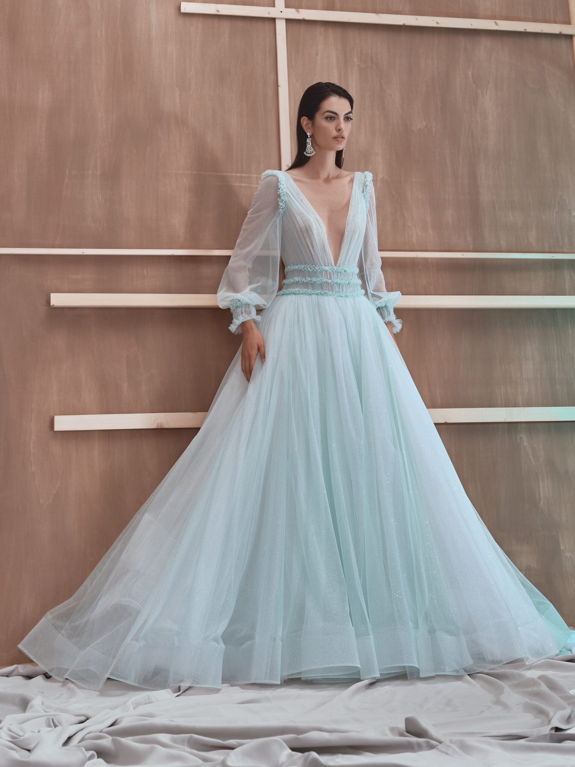 A woman stands in front of a wooden backdrop, wearing a voluminous light blue gown with sheer long sleeves, a deep V-neckline, and delicate detailing, with fabric pooling on the floor around her.
