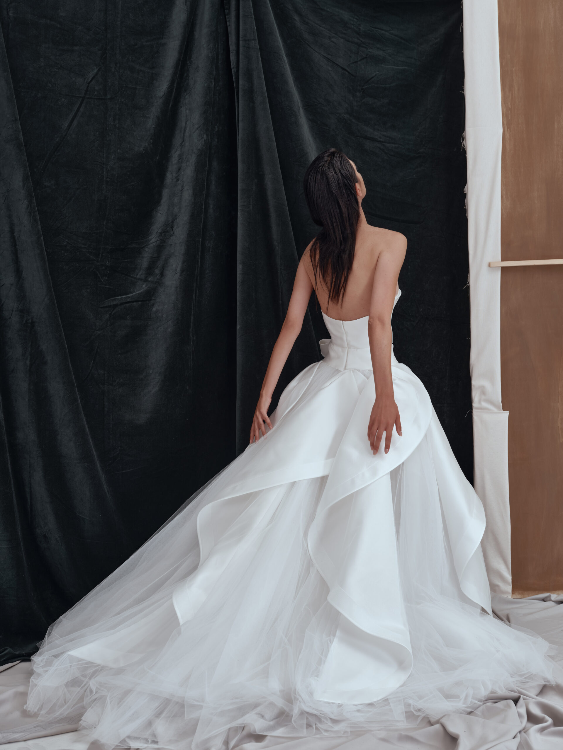 A woman in a strapless white wedding dress with a full, layered skirt stands with her back to the camera, posing against dark and neutral draped fabric backgrounds.
