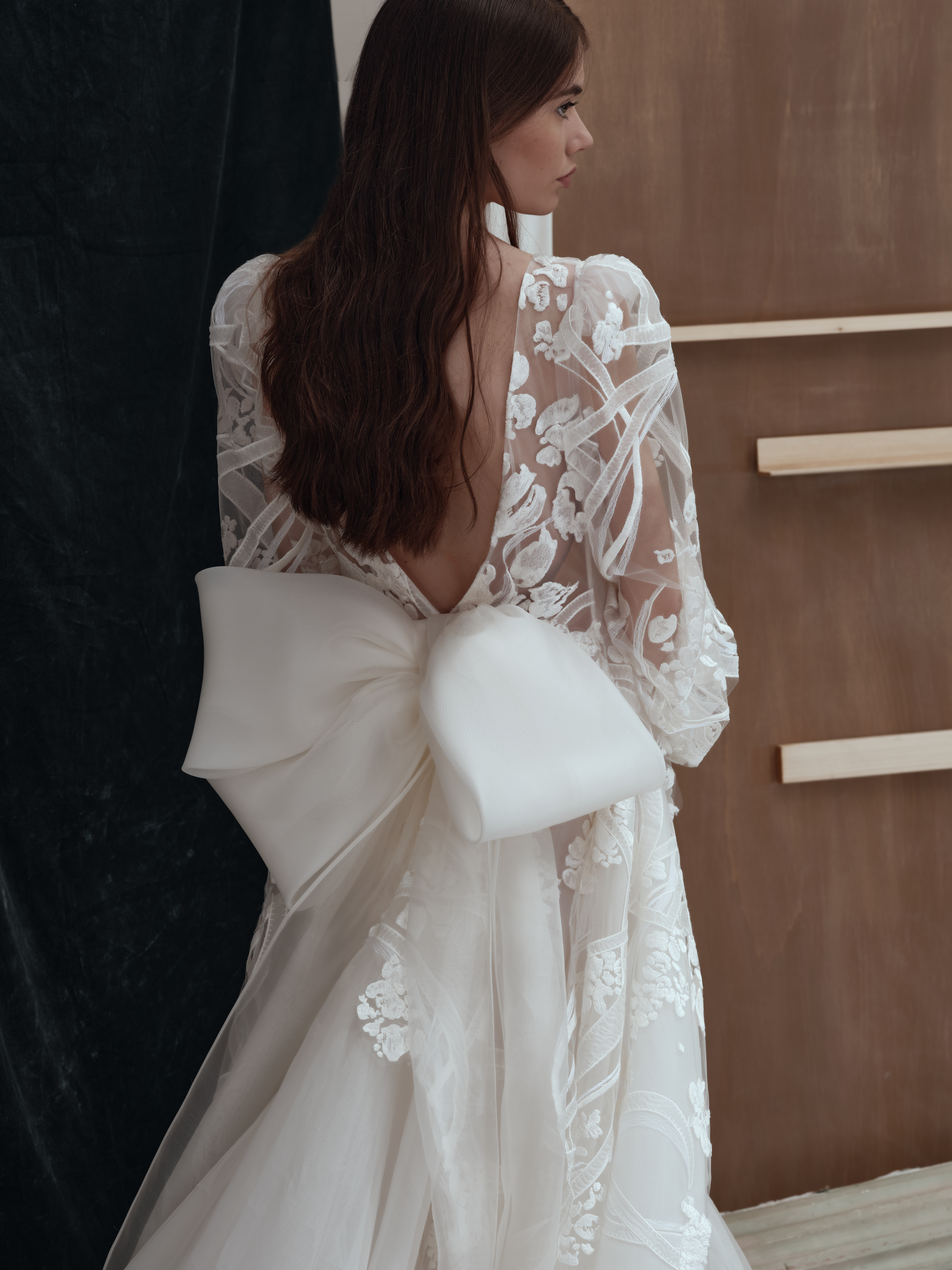 A woman with long brown hair wears a sheer, lace wedding dress with floral patterns and a deep V-back, featuring a large white bow at the lower back. She stands indoors against a dark and wooden background.