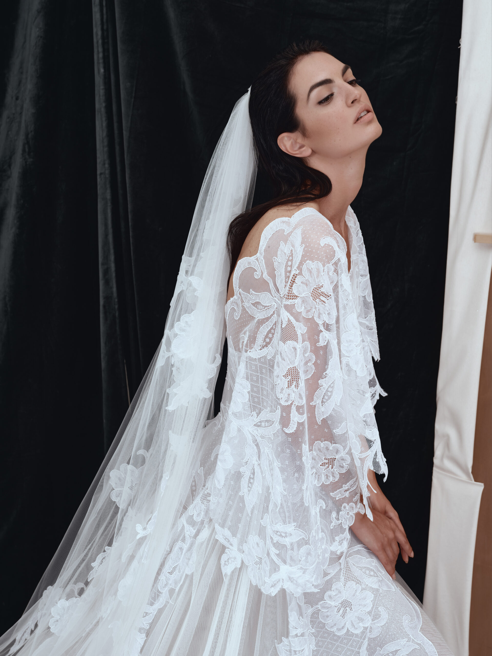A woman in a sheer, white lace wedding dress with floral patterns and a long veil poses with her eyes closed against a dark velvet backdrop.