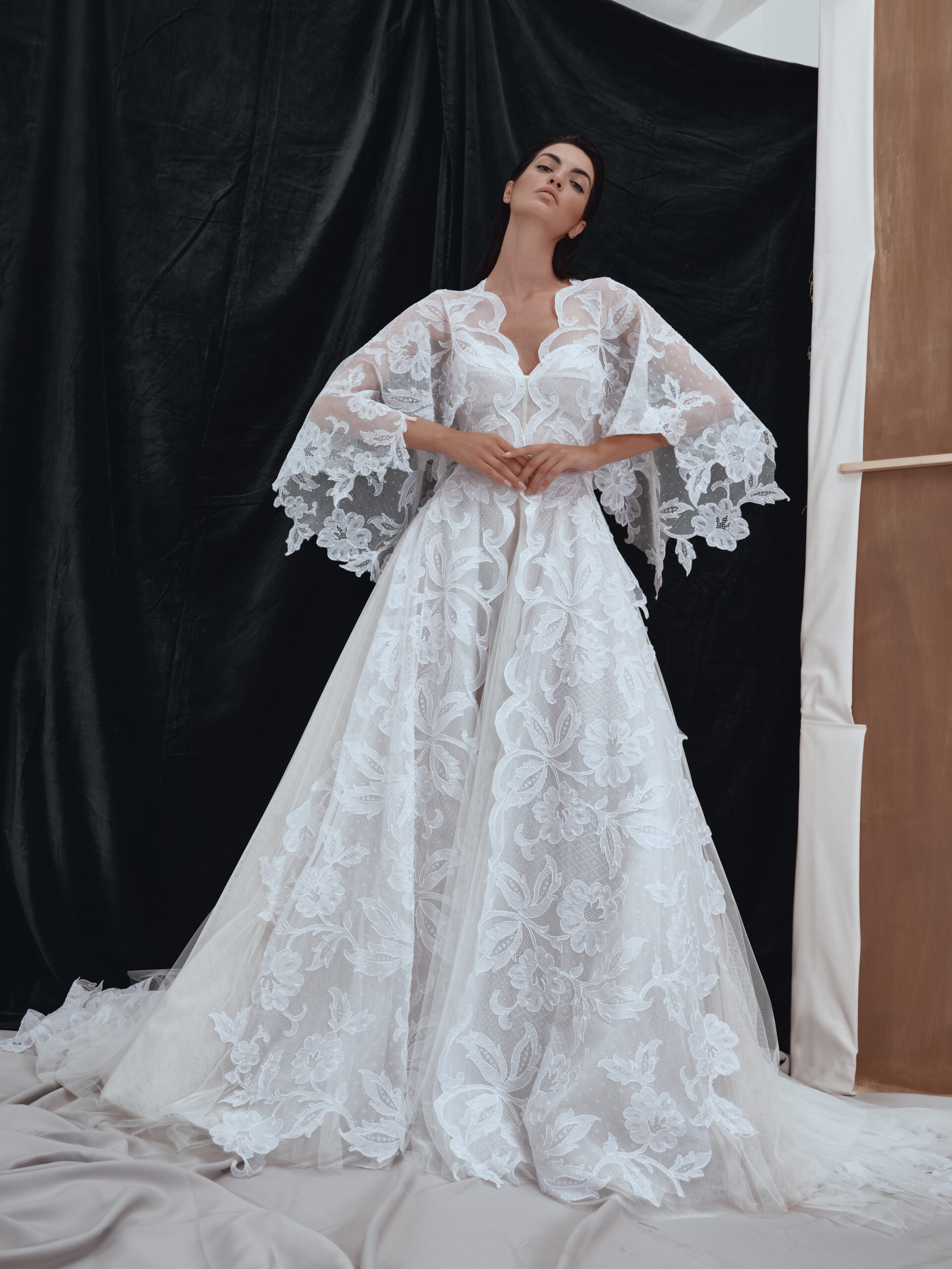 A woman wearing a flowing white lace gown with wide, sheer sleeves stands against a backdrop of black and beige fabrics, her hands clasped at her waist and her head tilted slightly back.