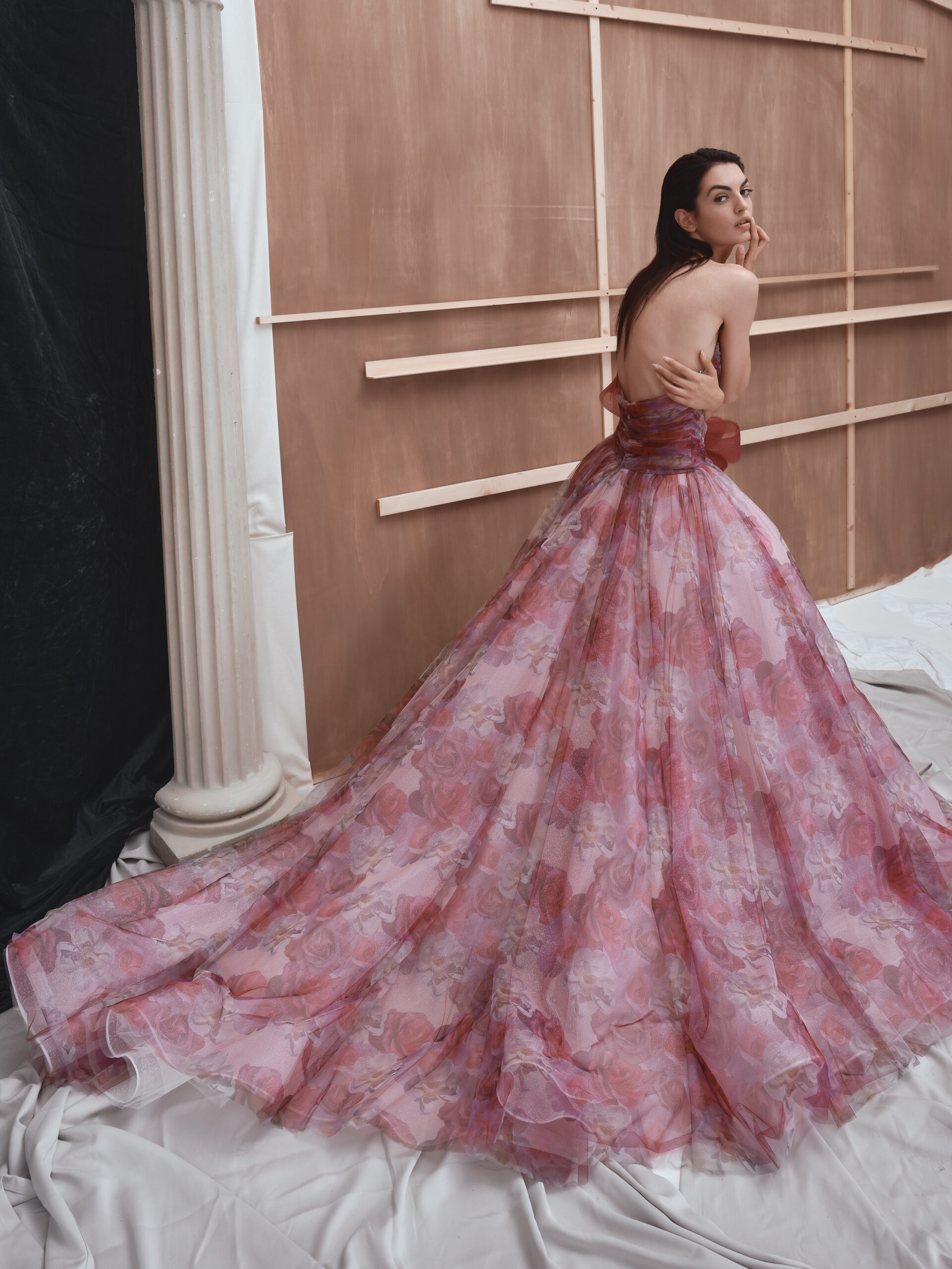 A woman poses in a backless, flowing pink floral gown with a long train, glancing over her shoulder. She stands in front of a wooden backdrop with a white draped floor and a tall column on the left.