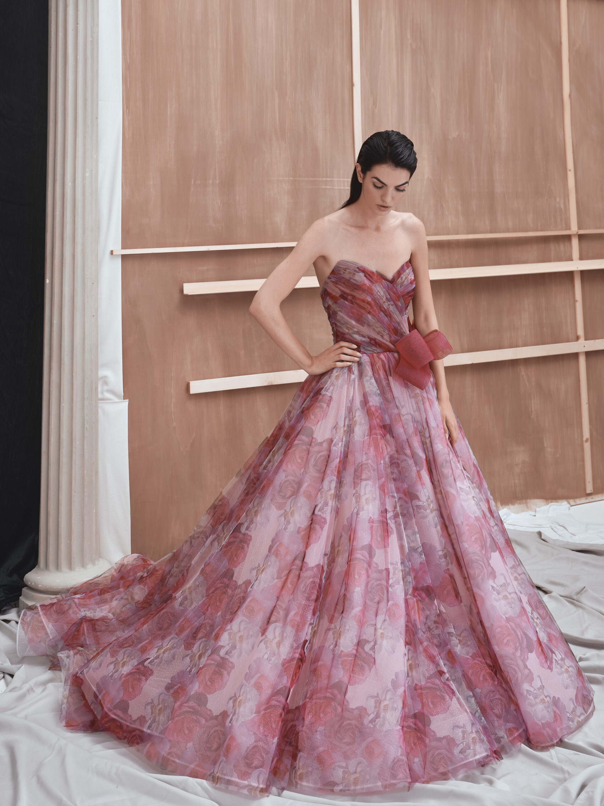 A woman in a strapless, floor-length pink and red floral gown stands indoors against a wooden backdrop, looking down with her hands resting on the dress.