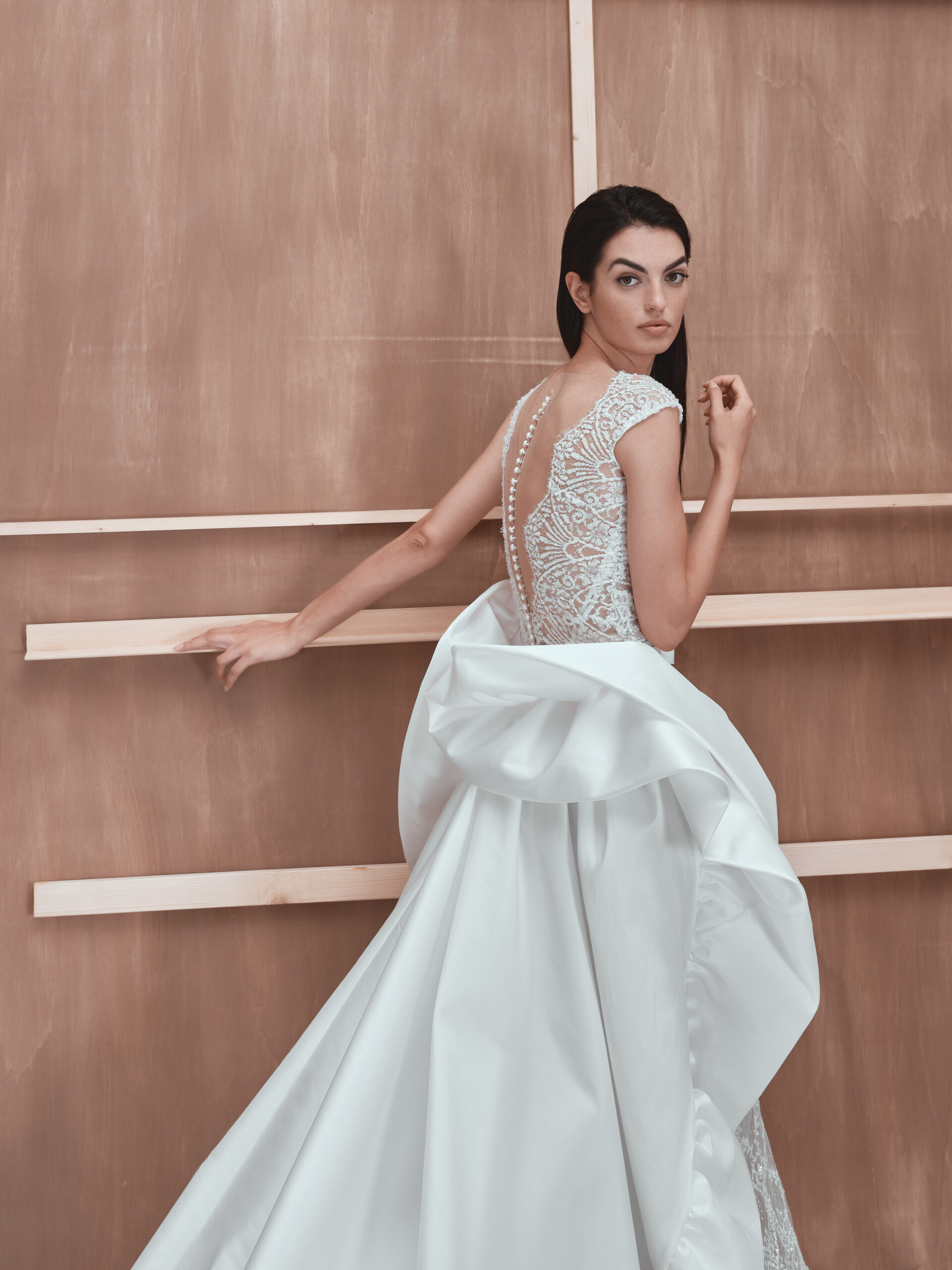 A woman in an elegant white wedding dress with lace detailing on the back and pearl buttons stands in front of a wooden backdrop, looking over her shoulder. The dress features a dramatic, voluminous train.