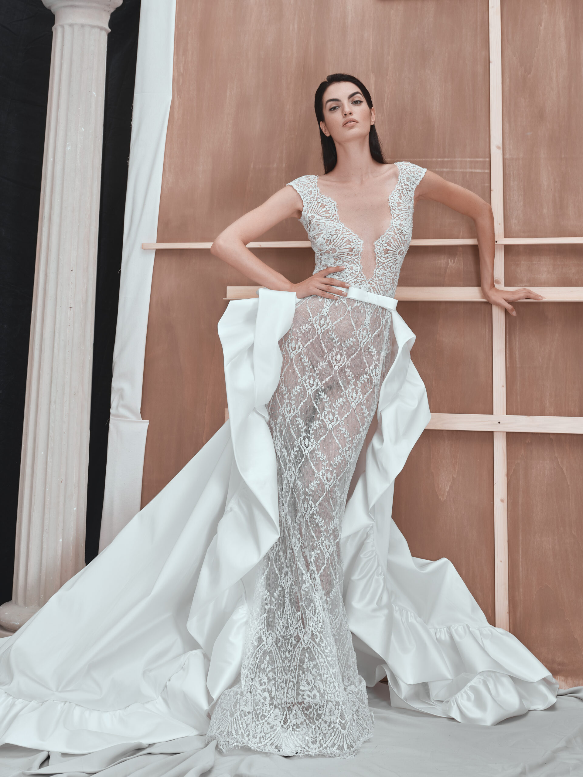 A woman poses in a sheer, lace, floor-length white gown with a dramatic ruffled train, standing against a wooden backdrop with a classical column to the side.