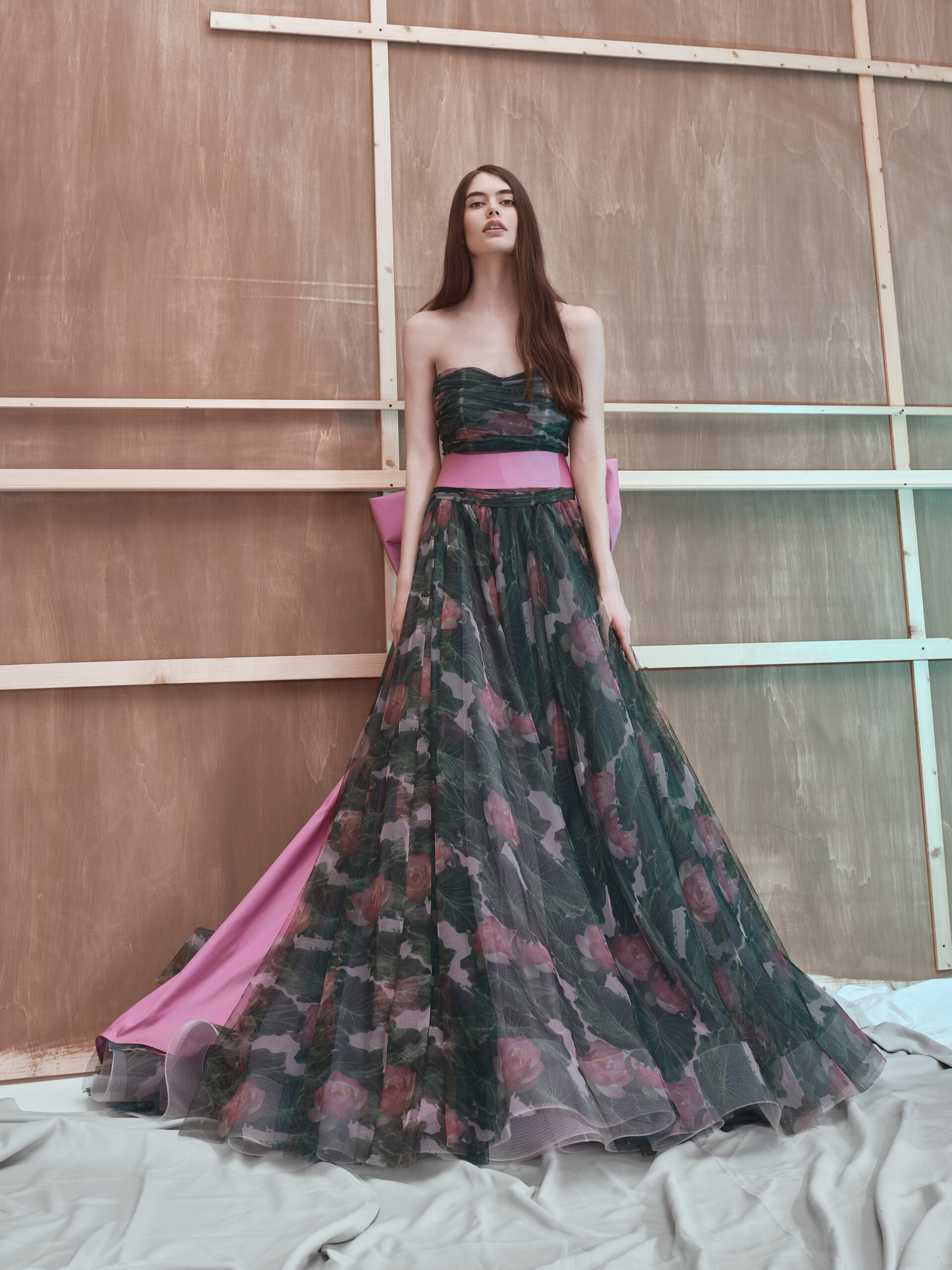A woman in a strapless, floor-length floral gown with a pink sash stands in front of a wooden panel backdrop, her long brown hair loose, and the gown’s tulle overlay drapes elegantly on the floor.