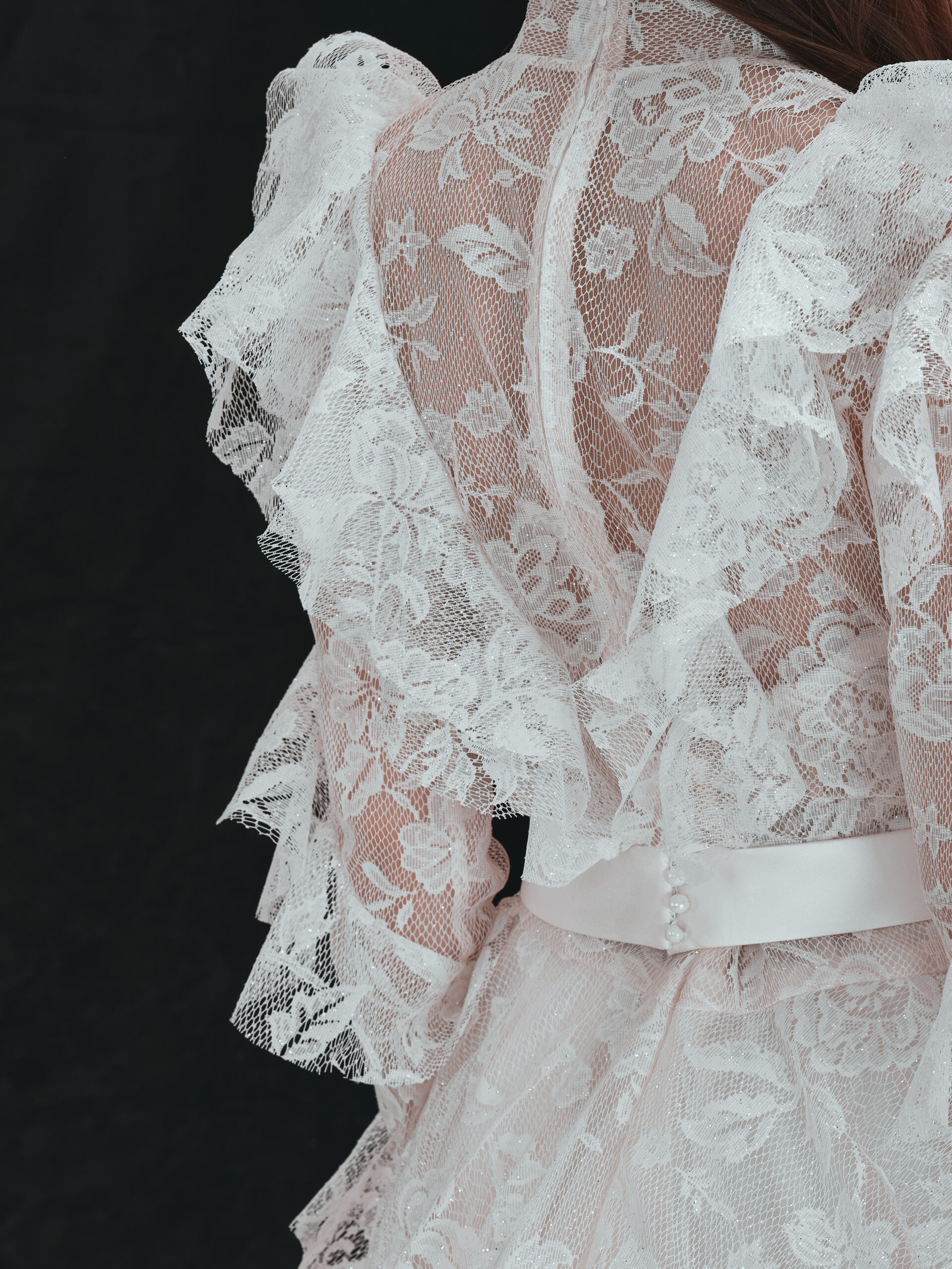 A close-up of the back of a person wearing a white lace dress with floral patterns, ruffled sleeves, and a satin waistband with small buttons. The background is dark, highlighting the intricate lace details.