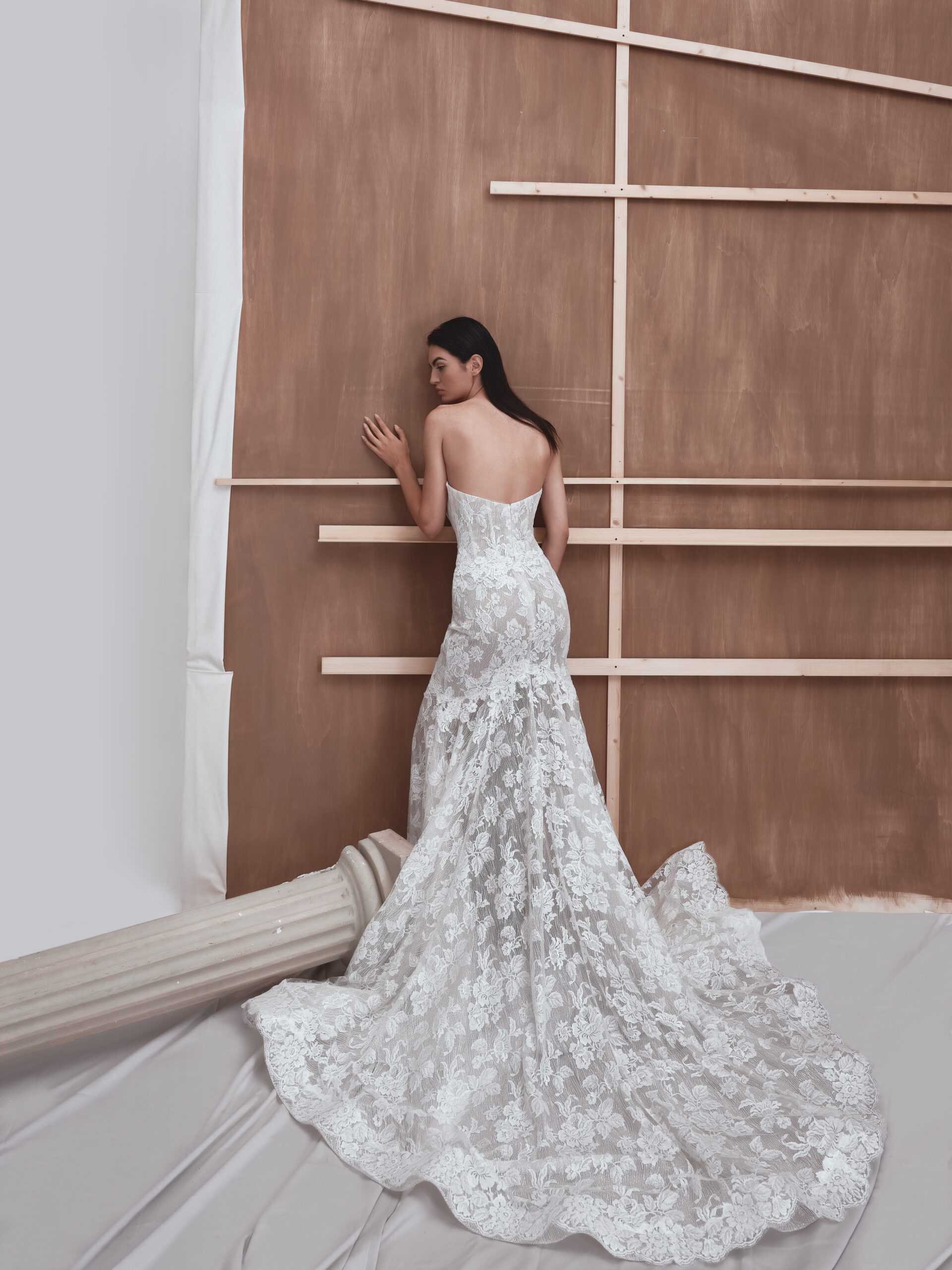 A woman in a strapless, white lace wedding gown with a long train stands with her back to the camera, facing a wooden backdrop. A fallen decorative column lies on the floor nearby.