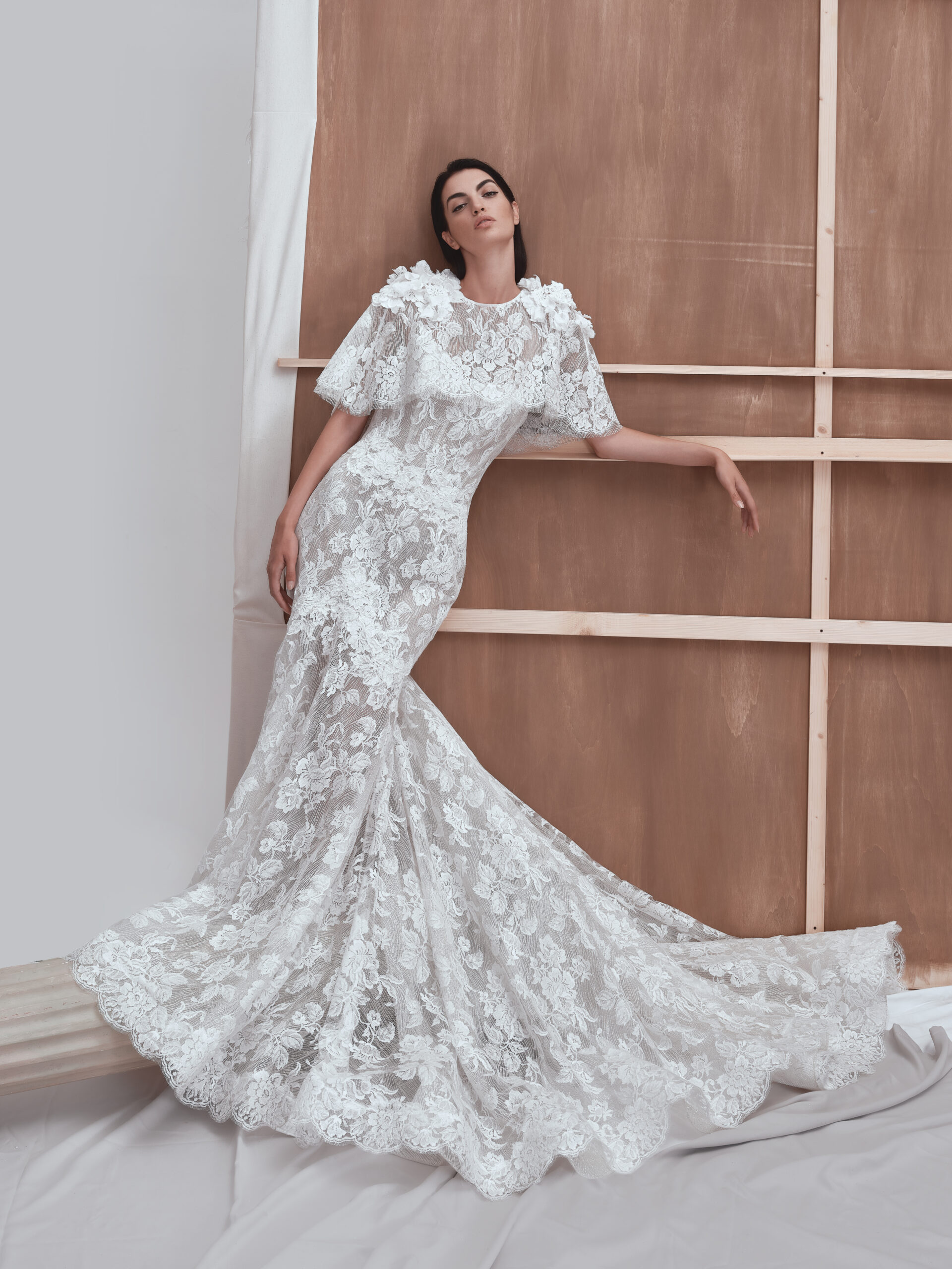A woman poses in a fitted, floor-length lace wedding gown with floral patterns and short flutter sleeves, leaning elegantly against a wooden backdrop.