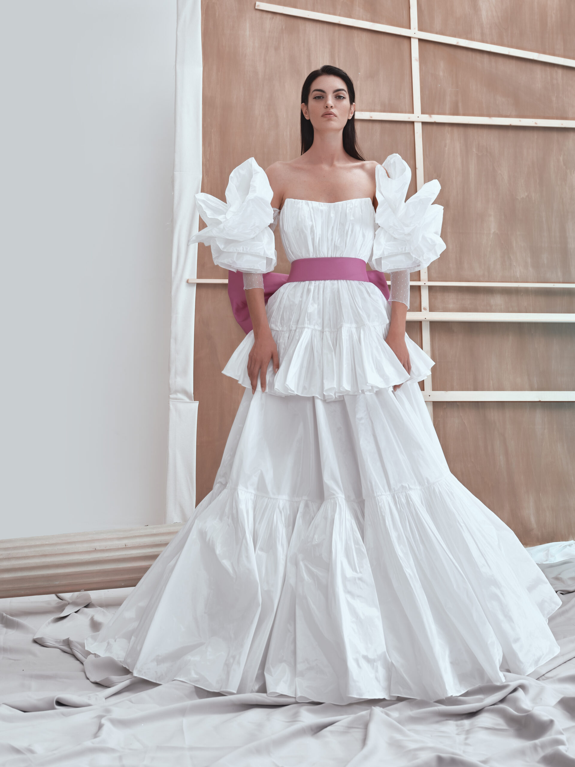 A woman stands in a white, ruffled off-shoulder gown with voluminous sleeves and a wide pink belt, posing indoors against a neutral backdrop with wooden slats and draped fabric on the floor.