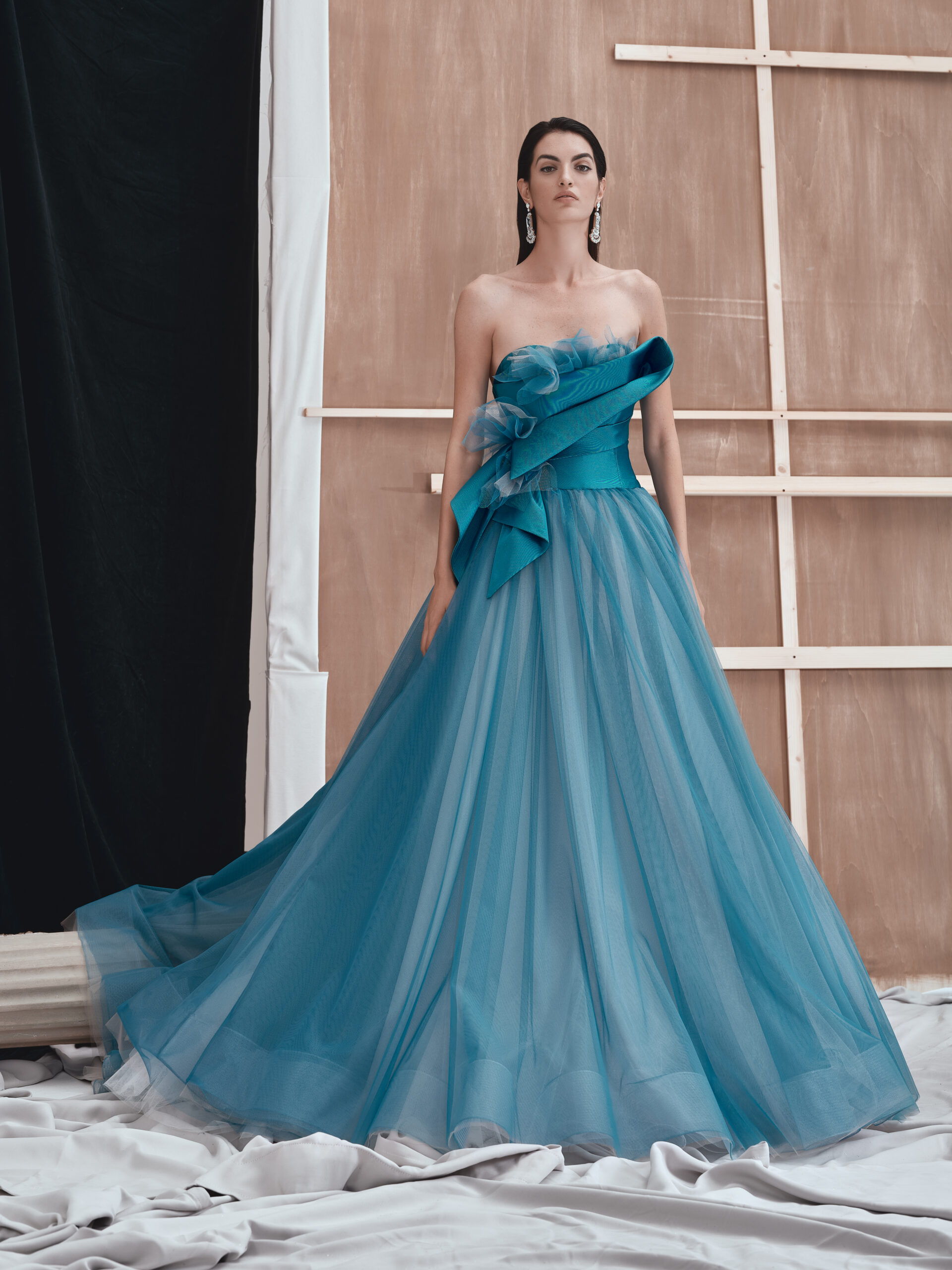 A woman in a strapless, flowing blue gown with a ruffled bodice stands in front of a wooden and fabric backdrop, looking confidently at the camera.