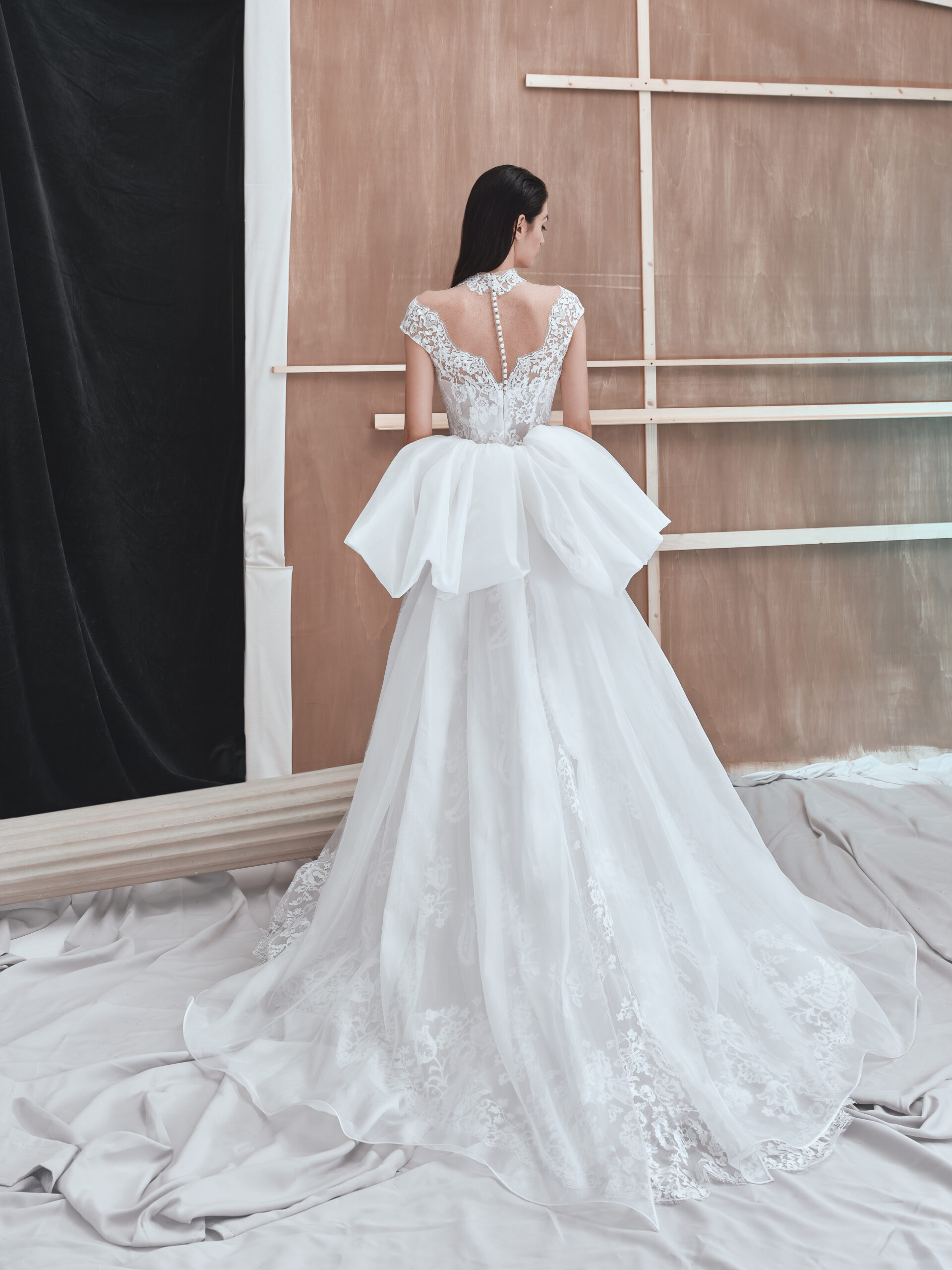A woman in a white, lace wedding gown with a large bow on the back stands facing away in a studio with wooden paneling and draped white fabric on the floor.