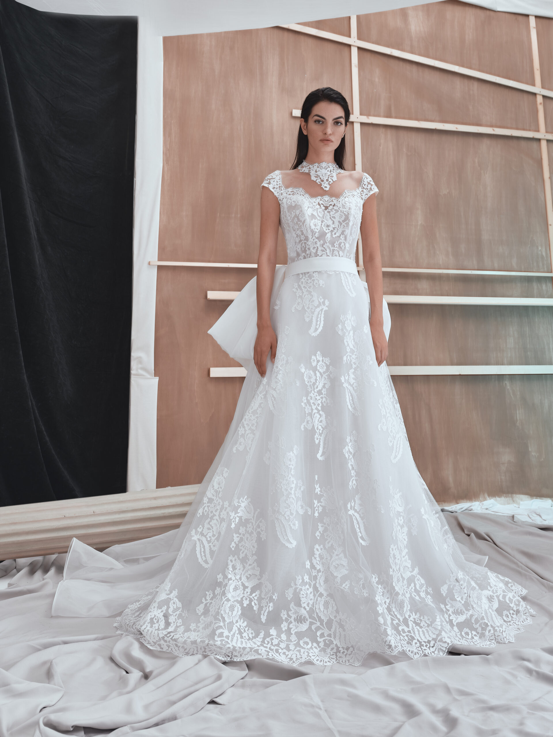A woman stands indoors wearing an elegant white lace wedding dress with cap sleeves and floral patterns, in front of a neutral, geometric wooden backdrop with draped fabric on the floor.