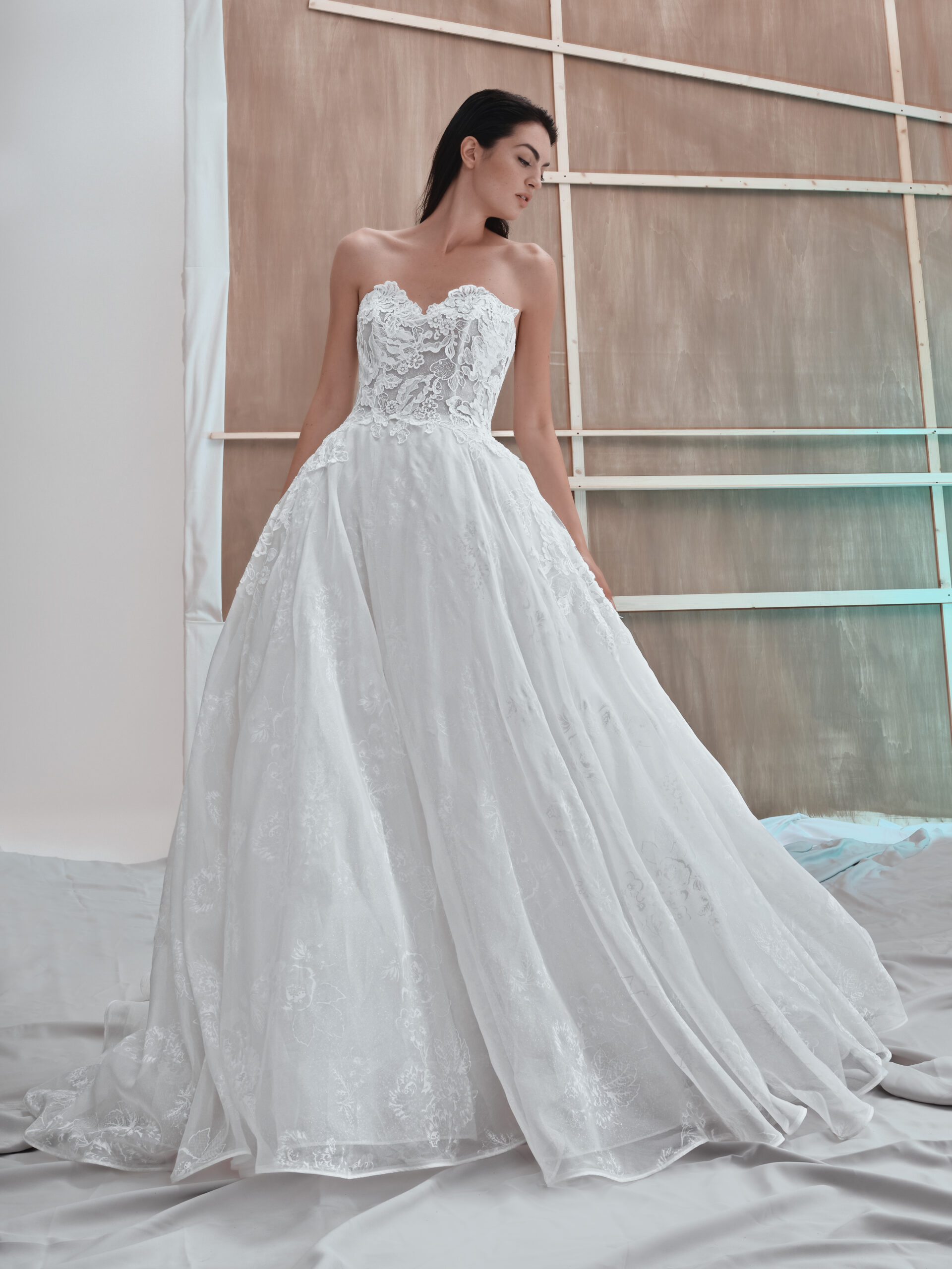 A woman in a strapless, lace-embellished white wedding gown stands against a minimalist, unfinished wooden backdrop, looking to the side. The dress features a fitted bodice and a full, flowing skirt.