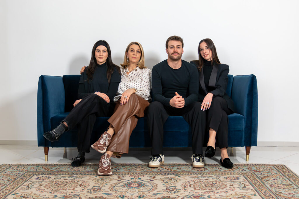 Four people sit on a blue sofa in a row, facing the camera. Three women and one man are dressed in stylish, semi-formal outfits. A patterned rug is on the floor in front of them, and the background is a plain white wall.