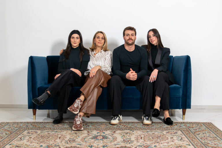 Four people sit on a blue sofa in a row, facing the camera. Three women and one man are dressed in stylish, semi-formal outfits. A patterned rug is on the floor in front of them, and the background is a plain white wall.