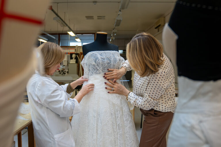 Two women adjust a white bridal gown on a black dress form in a workshop. One wears a white coat, the other a polka dot blouse, both focused on fitting the lace bodice of the dress.