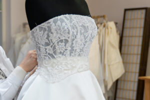 A person adjusts a white strapless wedding dress with a sequined lace bodice on a black mannequin in a tailoring studio. Fabric samples and a room divider are visible in the background.