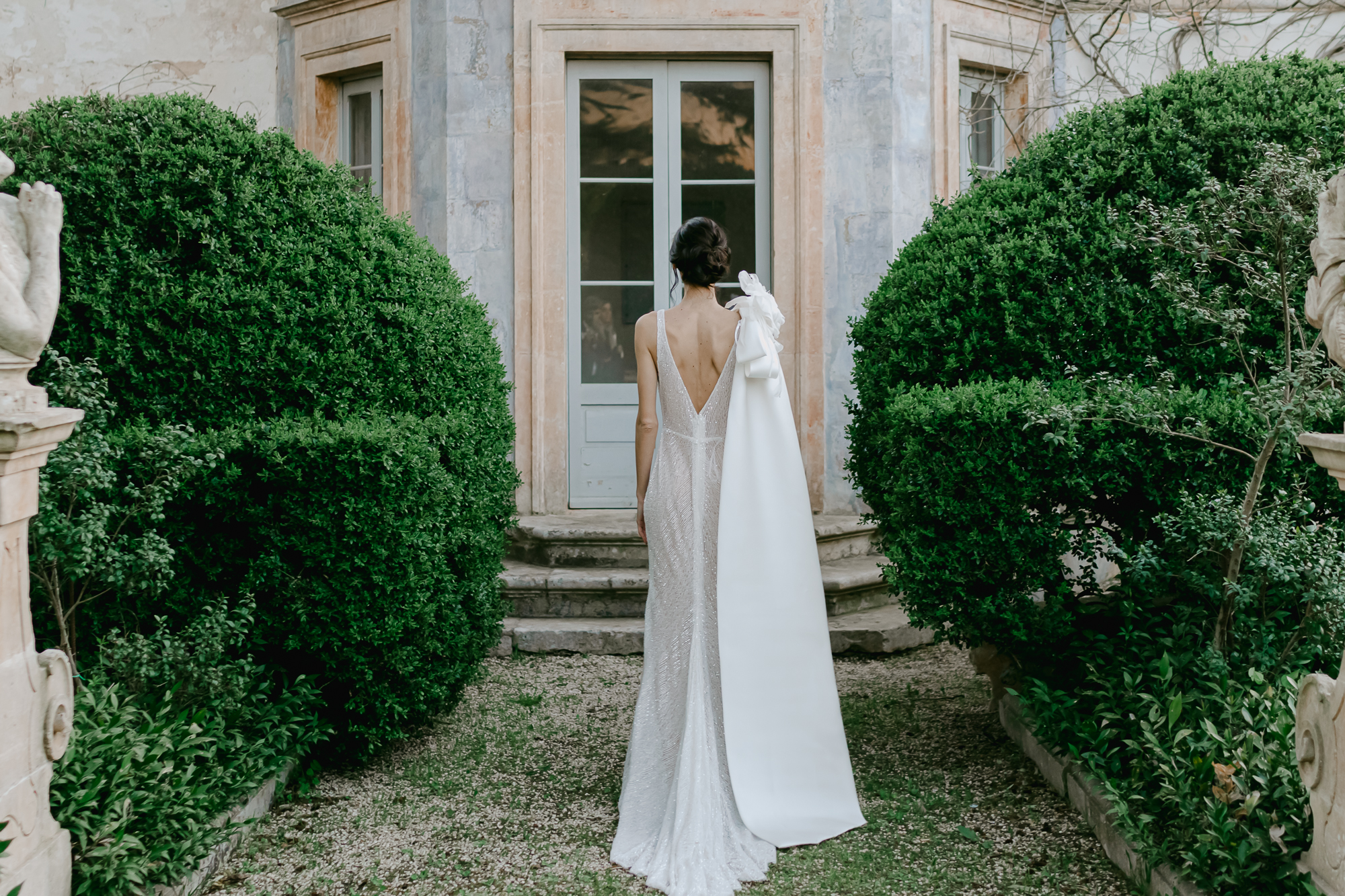 A woman in a white, backless gown with a long bow stands facing a closed door, surrounded by lush green bushes and stone architecture.