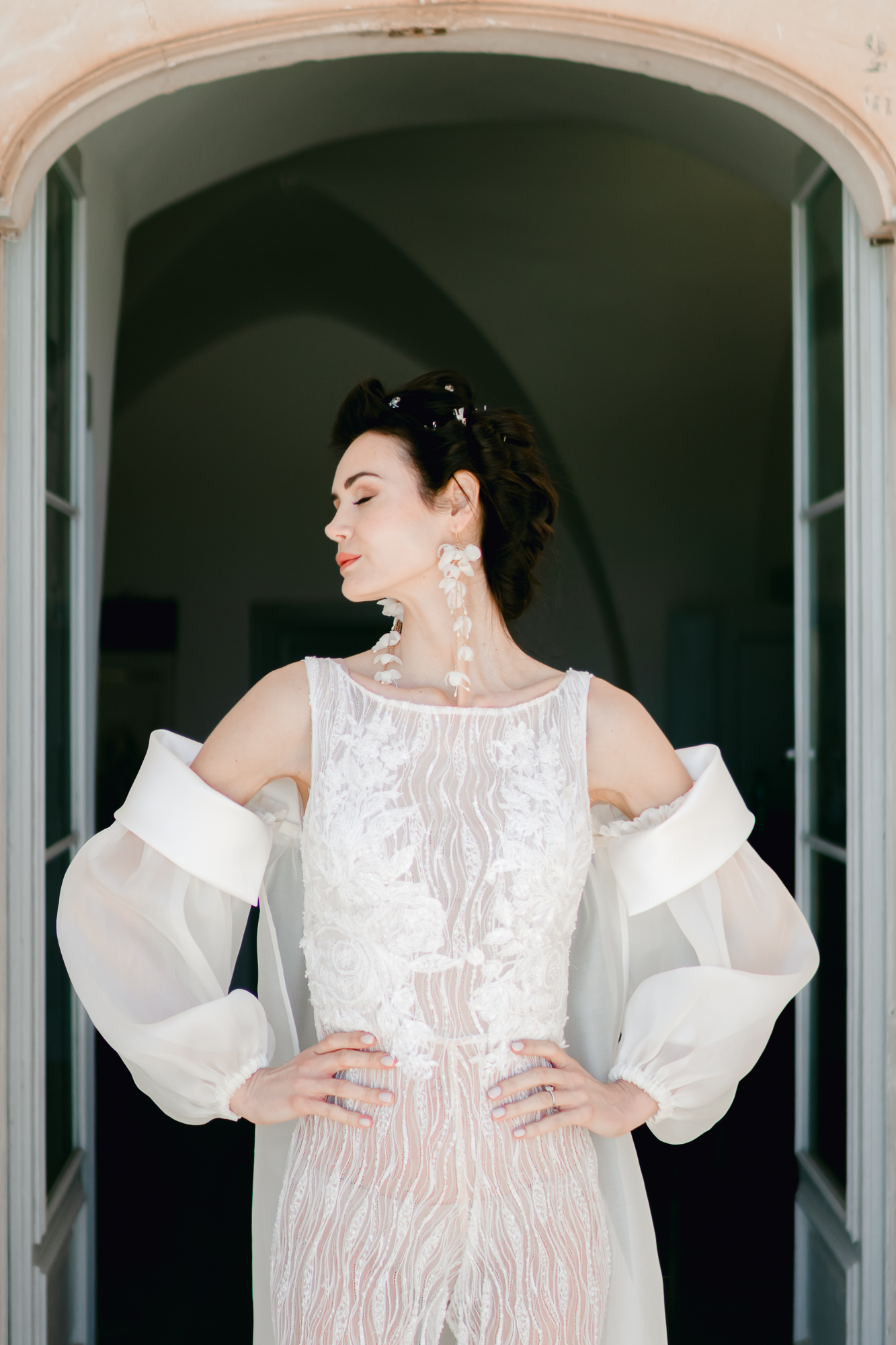 A woman in a white, sheer, lace wedding dress with dramatic, off-shoulder sleeves stands in a doorway, looking to the side with hands on hips and wearing statement earrings.
