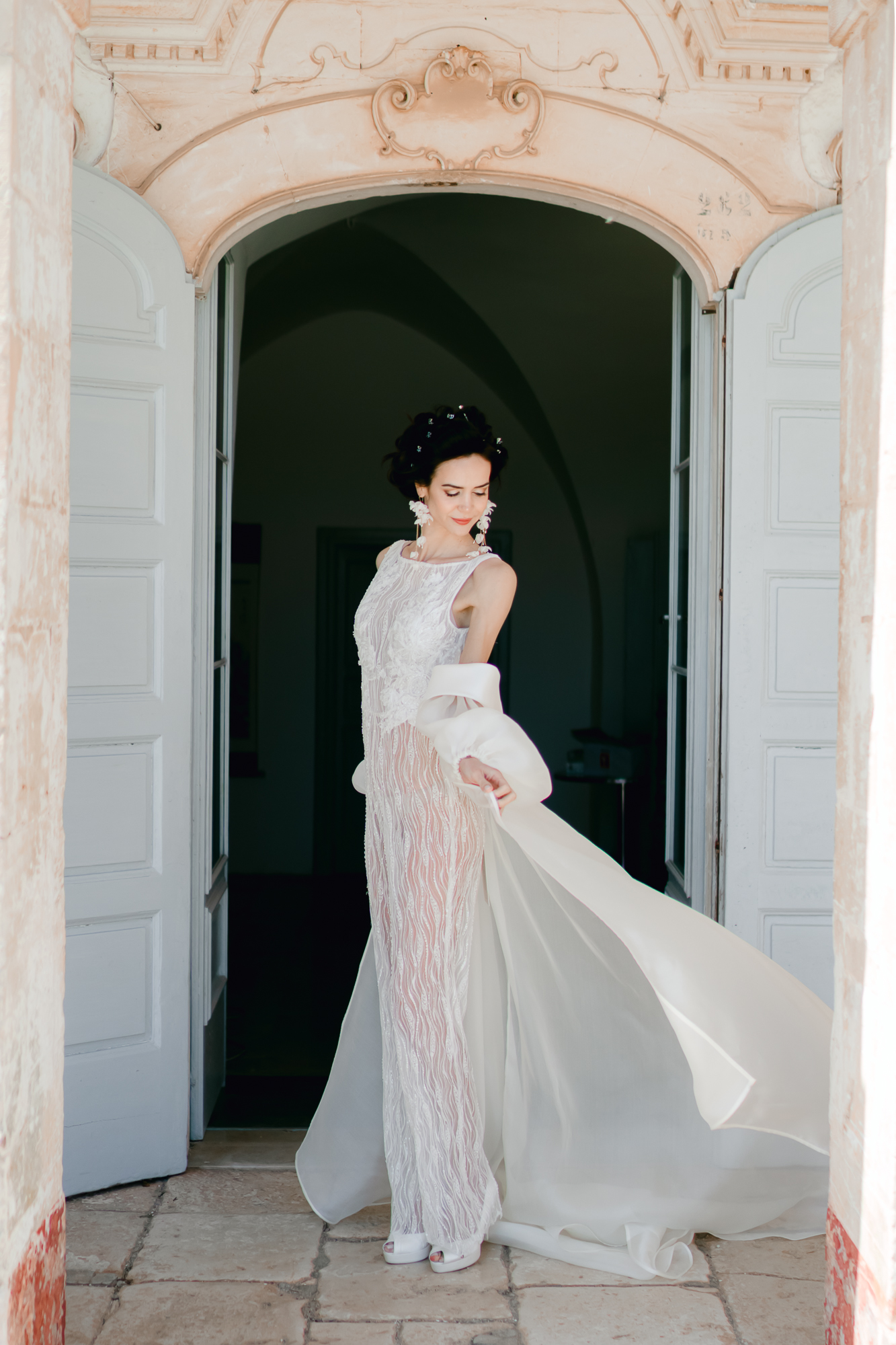 A woman in a white, sheer wedding dress stands in a doorway with arched stonework, gracefully holding part of her flowing gown, looking down with an elegant pose.